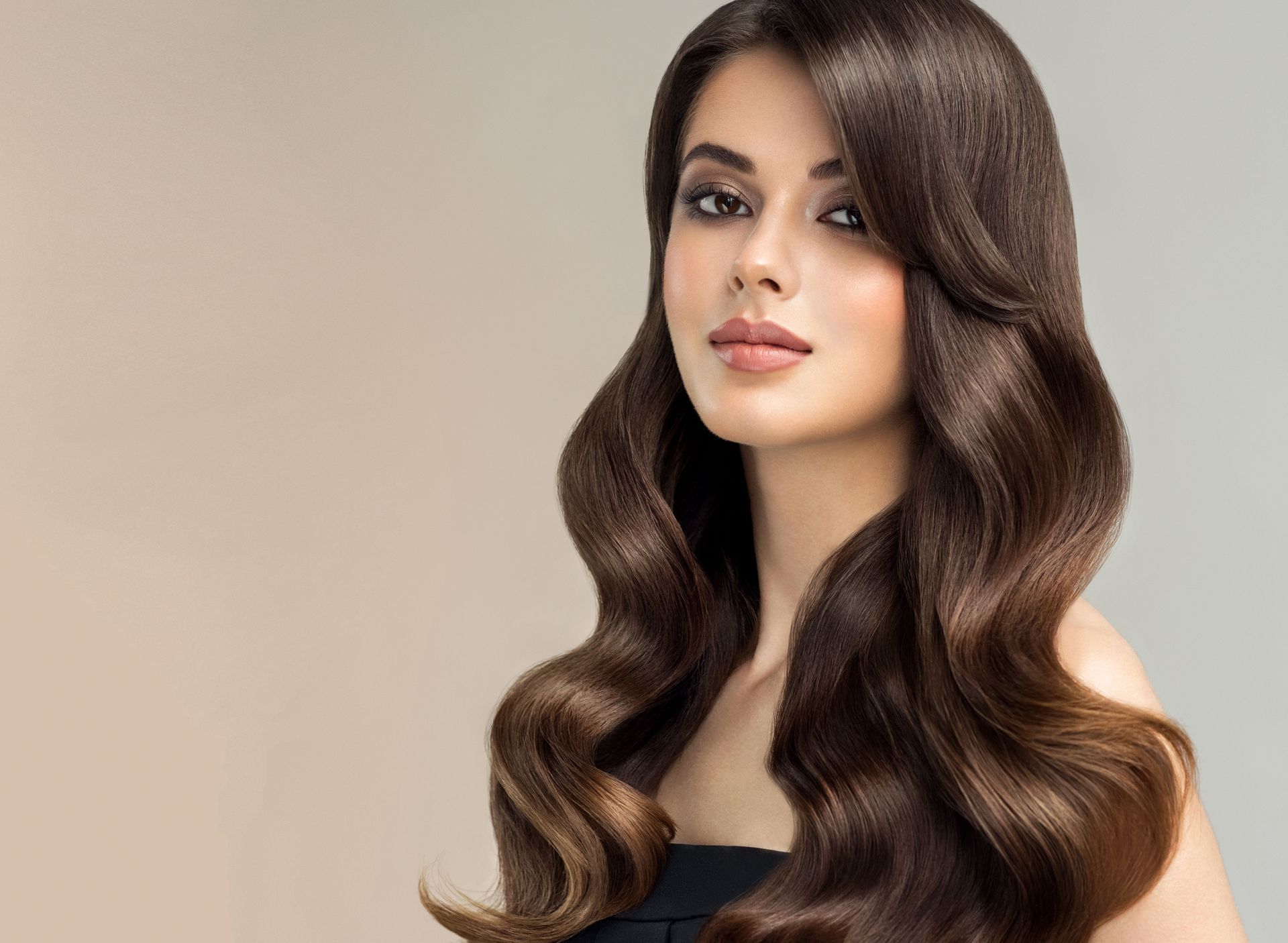 Woman with long, wavy brown hair, looking towards the camera, against a light background.