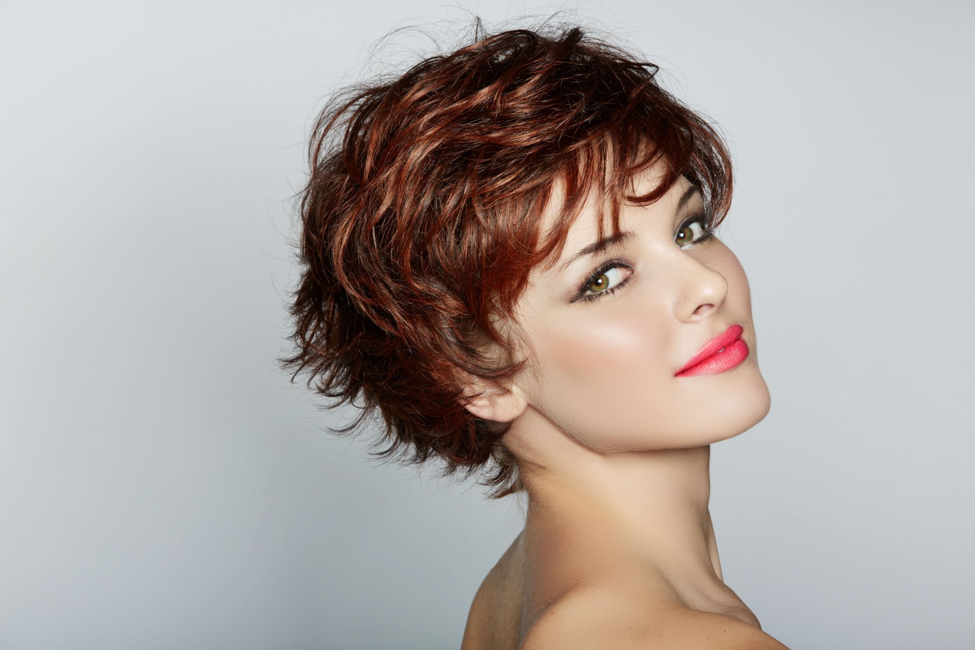 Woman with red, tousled short hair and bright pink lipstick, looking over shoulder.