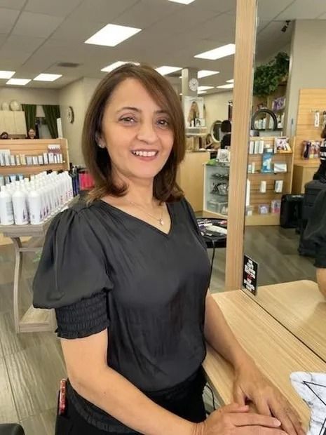 Woman in a salon, smiling, wearing a black top. Hair is styled and she stands near a mirror.