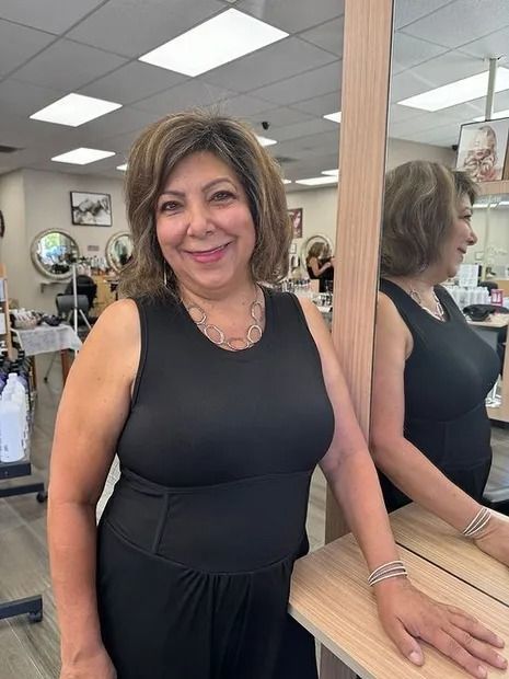 Woman with short brown hair smiles, stands by mirror in salon, wearing black dress.