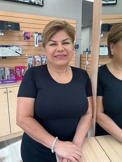 Woman with short, light brown hair, wearing a black shirt, smiles in a shop setting, with a mirror visible.