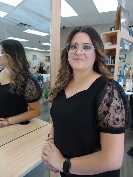 Woman with glasses in a salon, wearing a black top with lace sleeves, looking at the camera with arms crossed.
