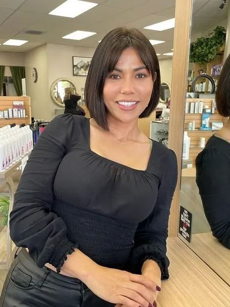 Woman with short, dark bob smiling in a salon, wearing black top and pants, leaning on a counter.