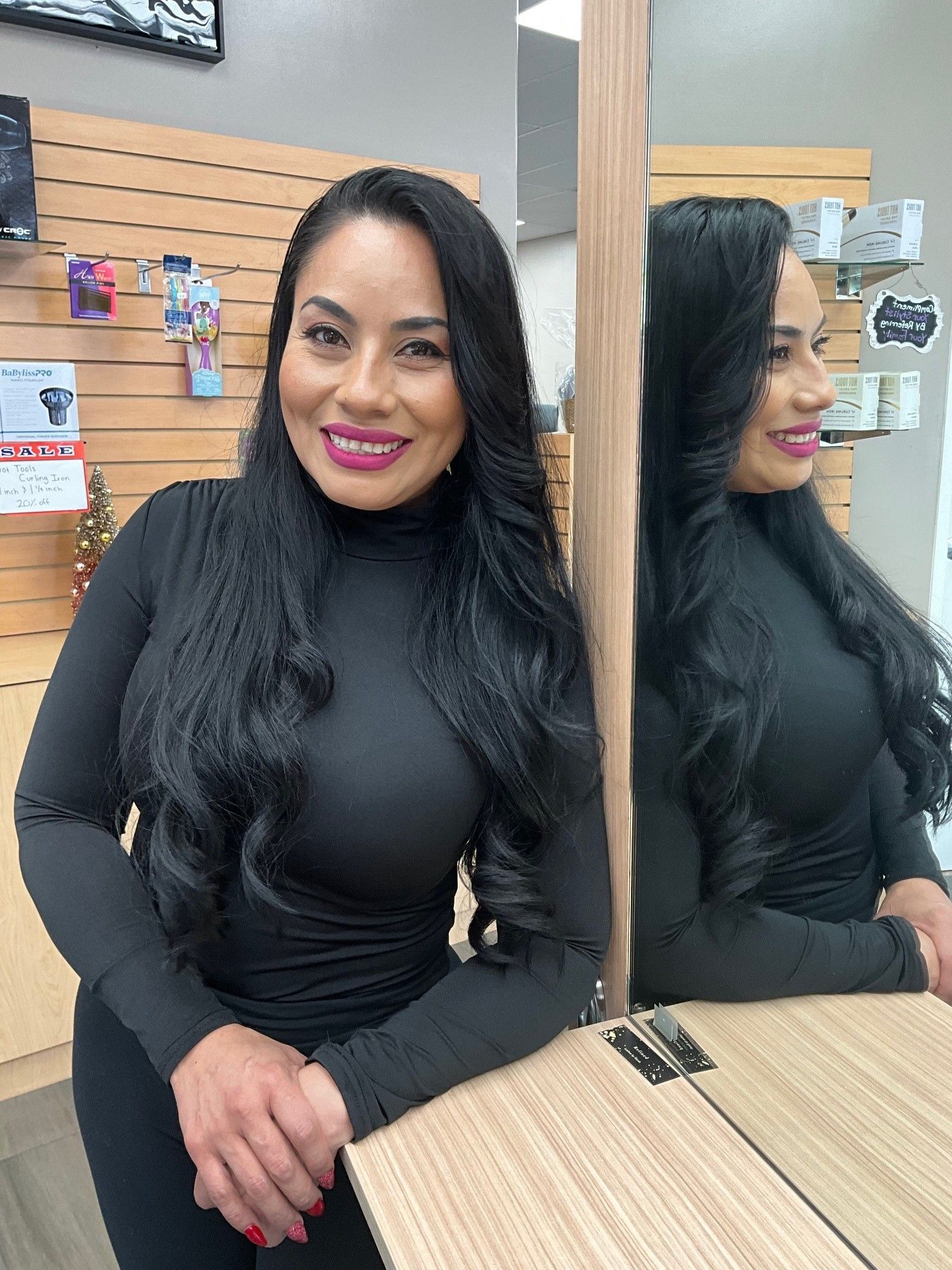 Woman smiling, sitting in salon chair, wearing black top and jacket, dark hair with layers.