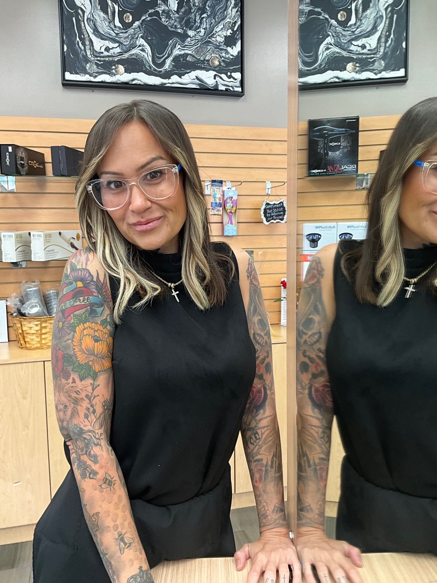Woman with dark hair, blonde highlights, black outfit, smiling, in a salon, leaning near a mirror.