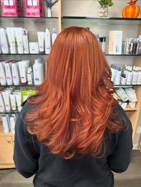 Person with long, vibrant red hair, styled in loose waves, stands in front of a shelf of beauty products.