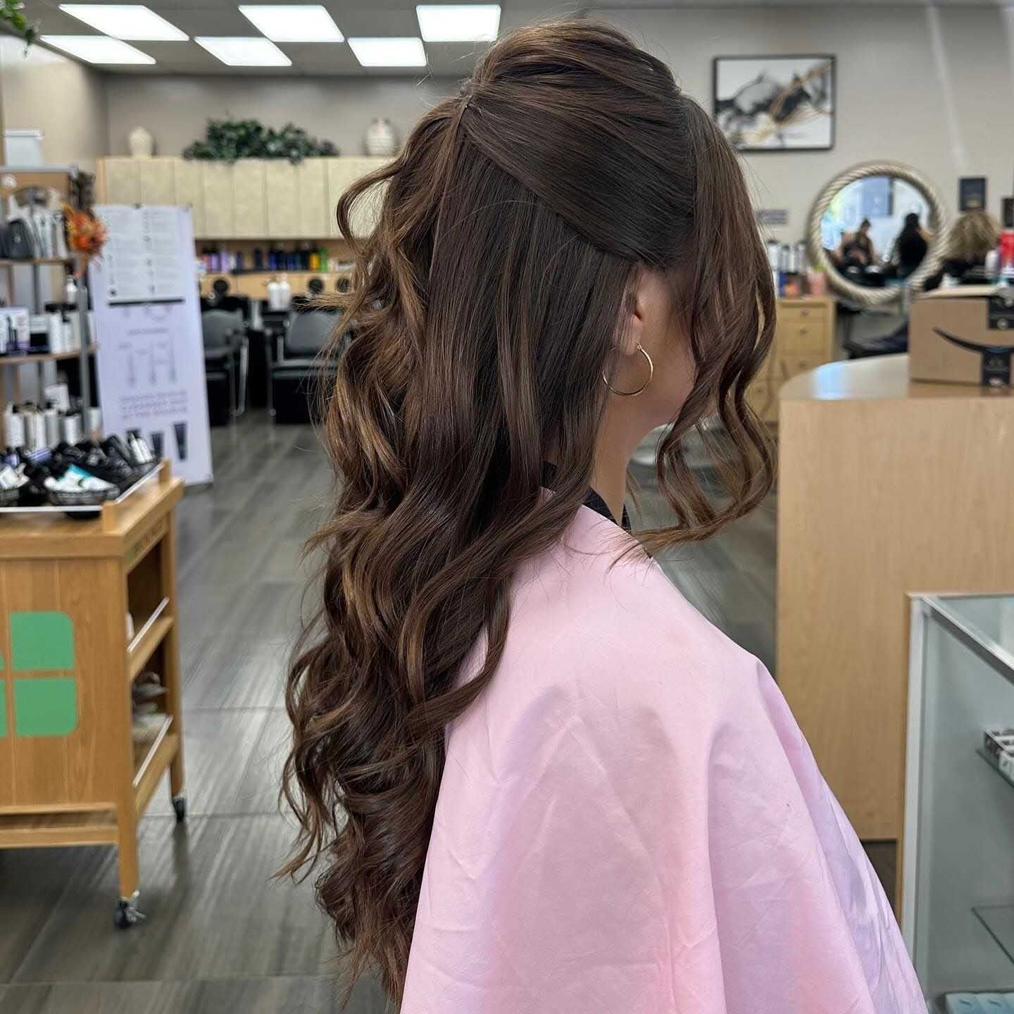 Woman with brown hair styled in a half-up, half-down curly hairstyle, in a salon, wearing a pink smock.