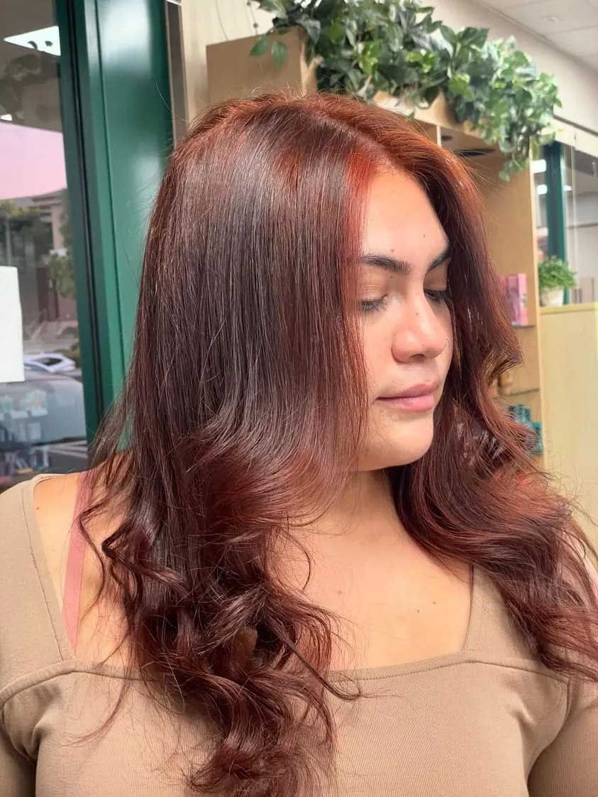 Woman with reddish-brown hair styled in waves and layers; she wears a neutral top and is in a salon.