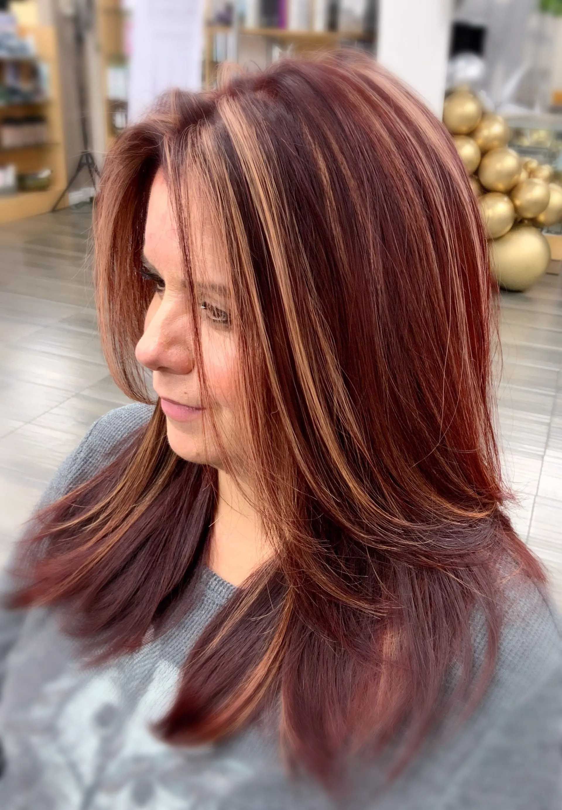Woman with layered auburn hair with blonde highlights; inside a salon.