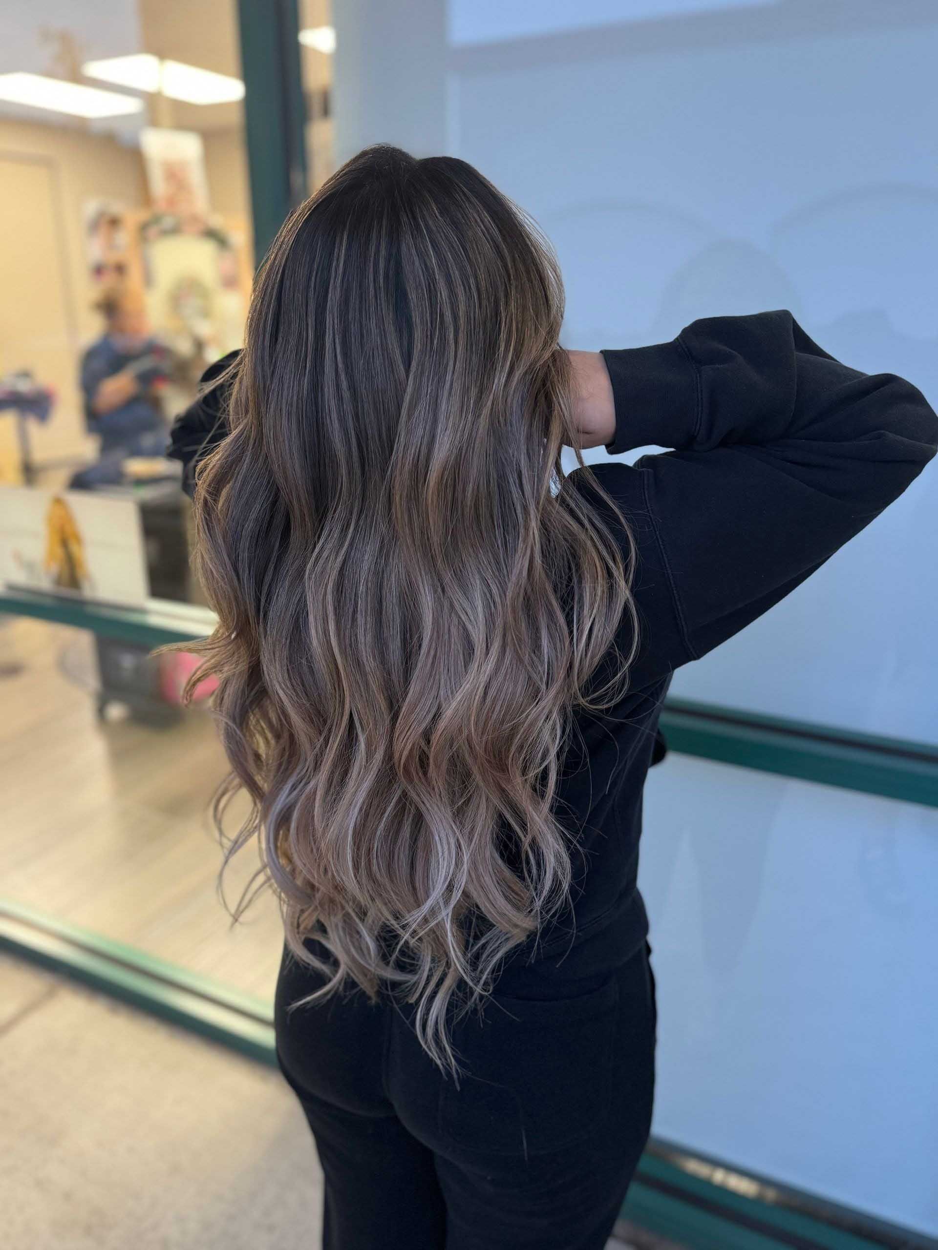 Person with long, wavy, dark-to-light brown hair in front of a window. Back to camera, hands on head, wearing black clothing.