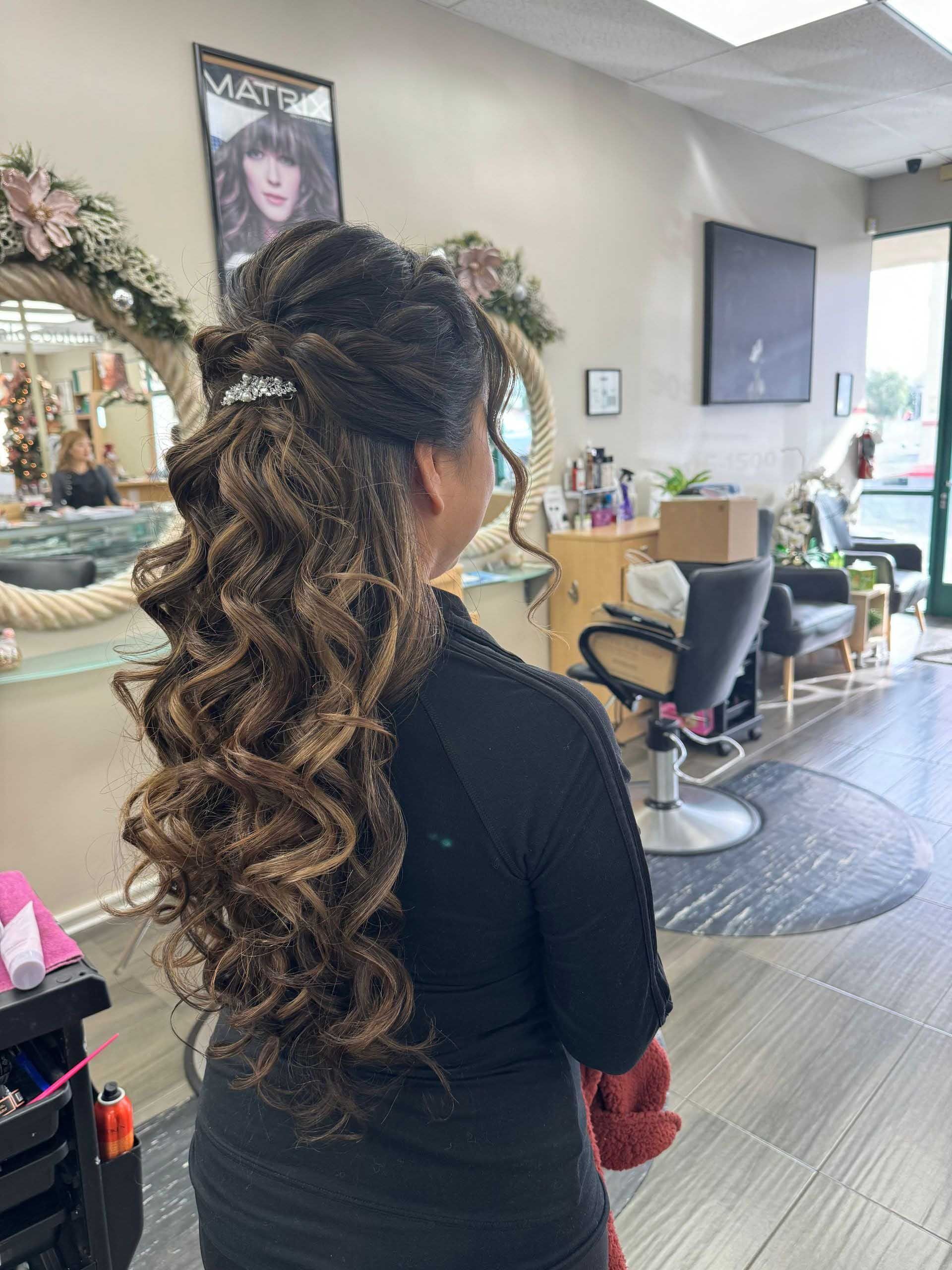 Woman with styled brown hair in a salon. Long curls cascade from an updo, in front of a mirror and other salon elements.