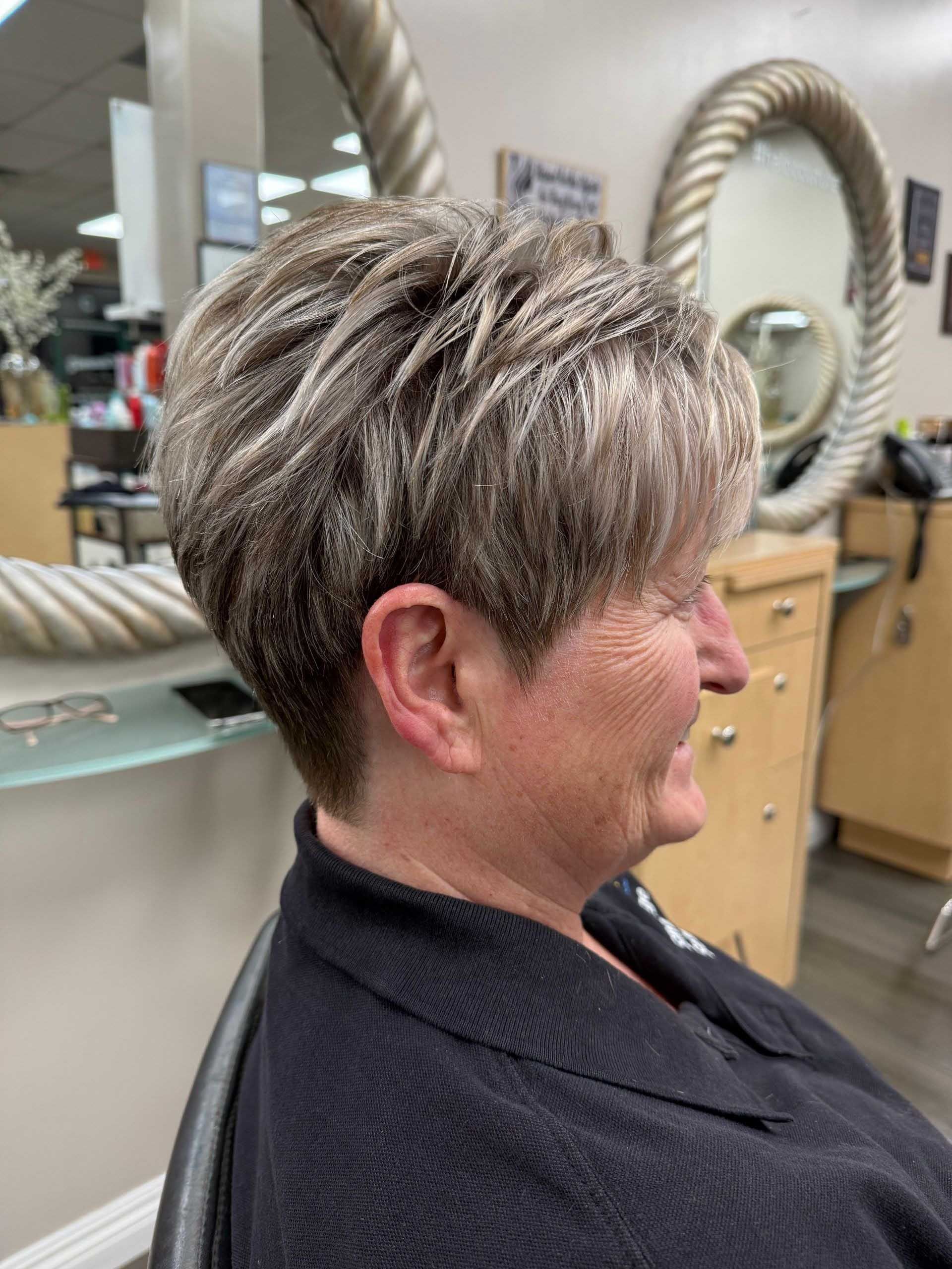 Woman with a short, layered hairstyle, in a salon setting, smiling. Hair is highlighted.
