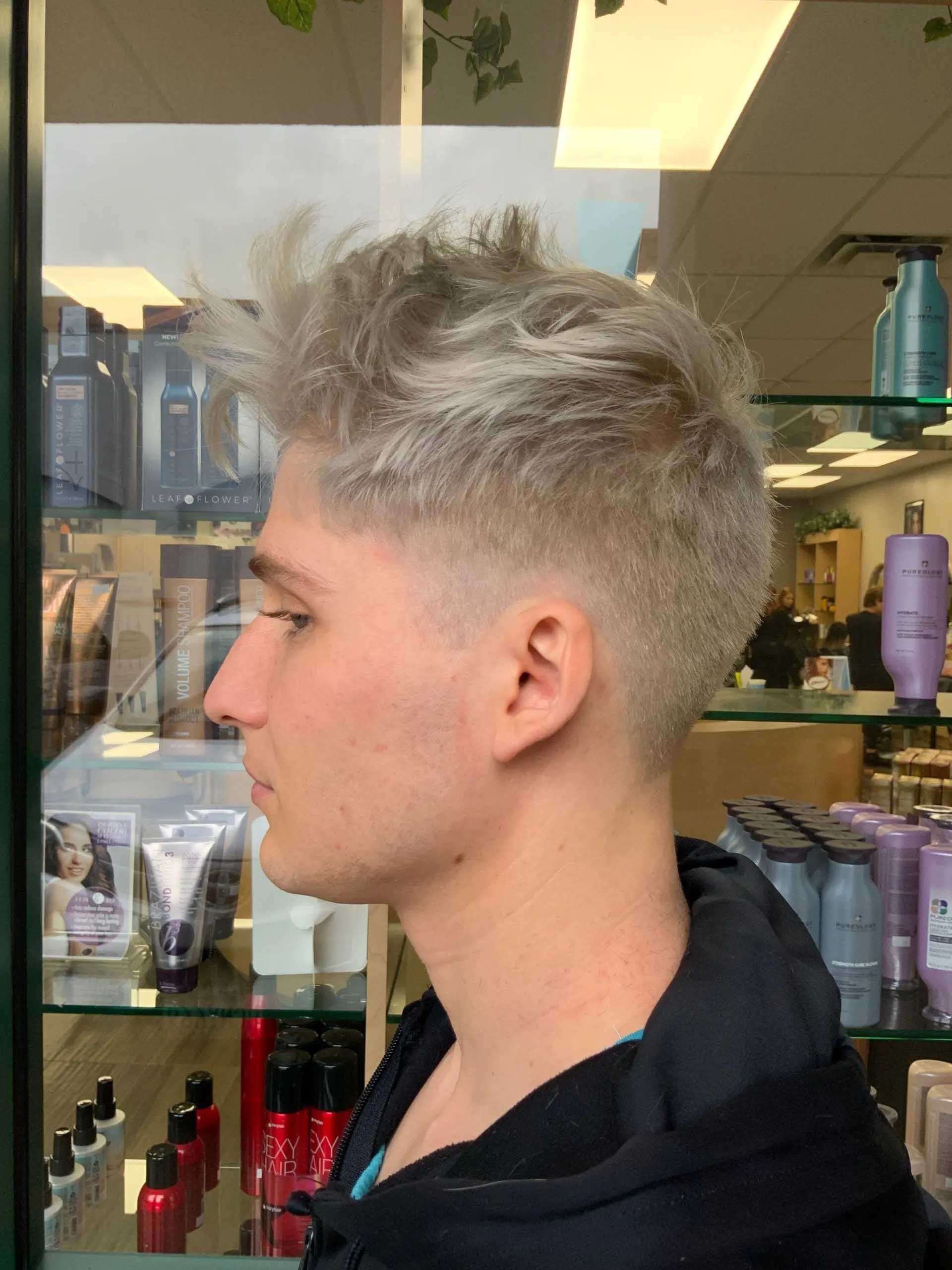 Man with light-colored, spiky hair, wearing a dark jacket, in a hair salon.