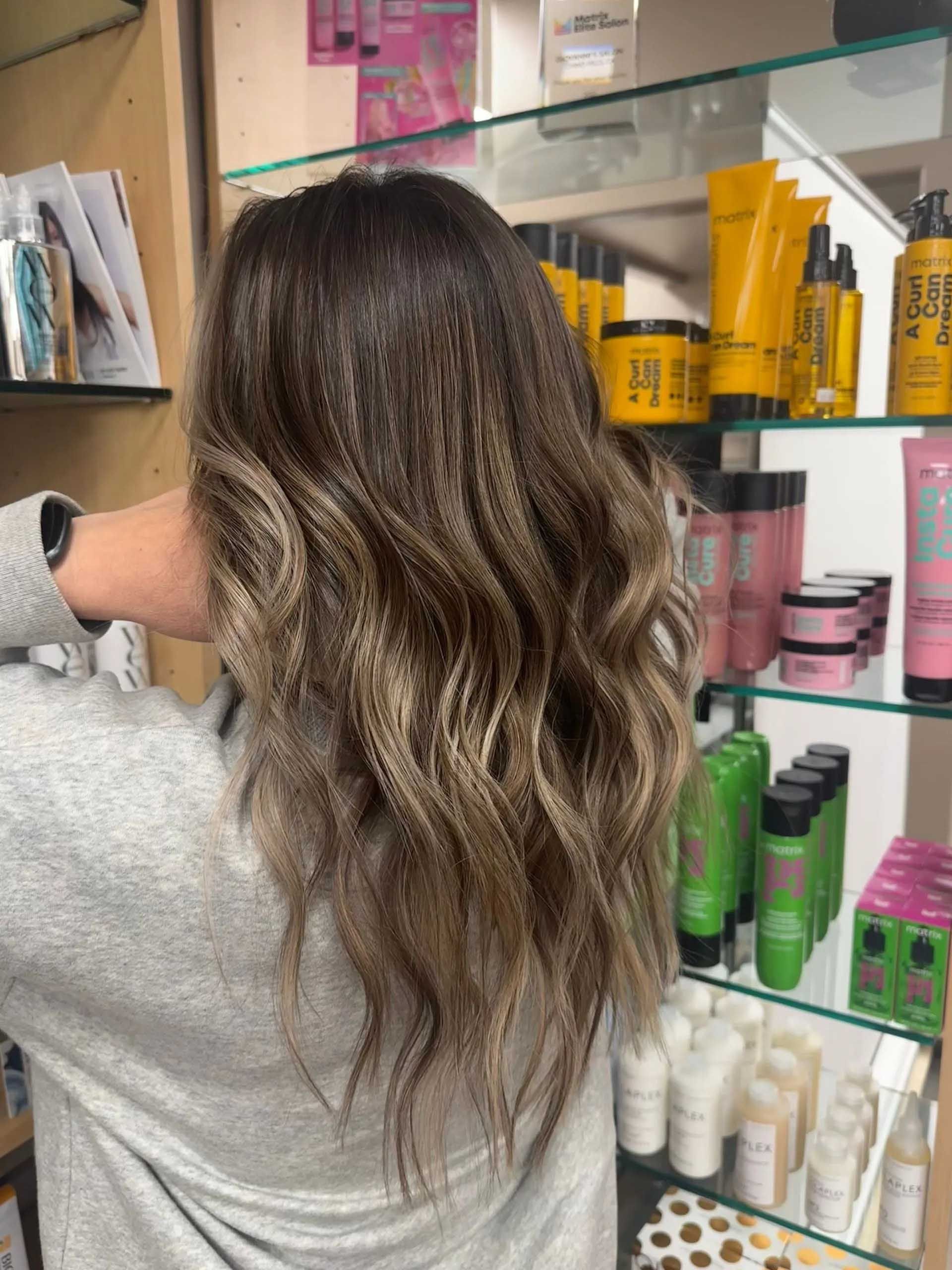 Woman with wavy brown and blonde hair, in front of hair product display.