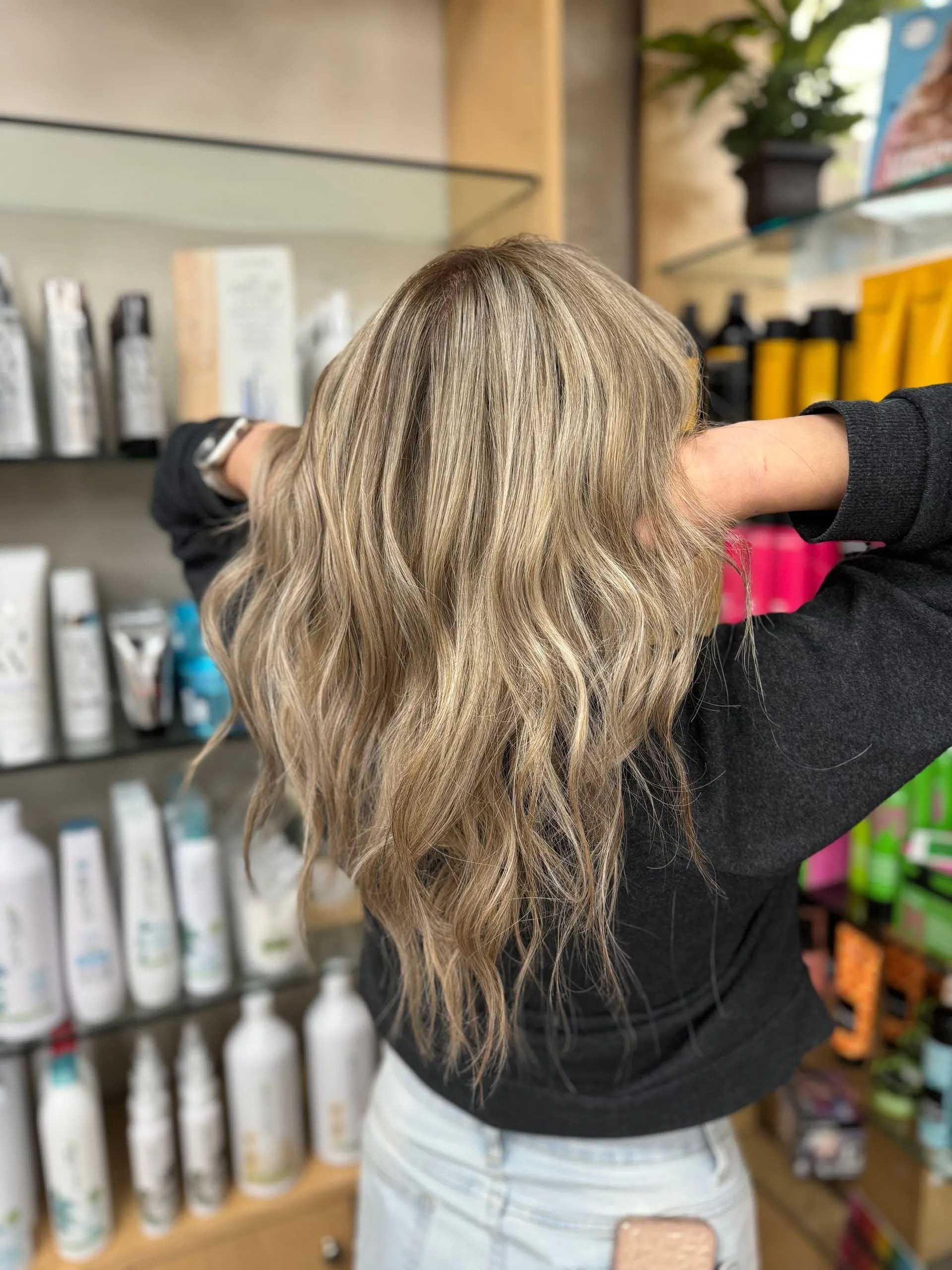 Woman with light brown crimped hair at a salon, holding hands behind head, in front of product shelves.