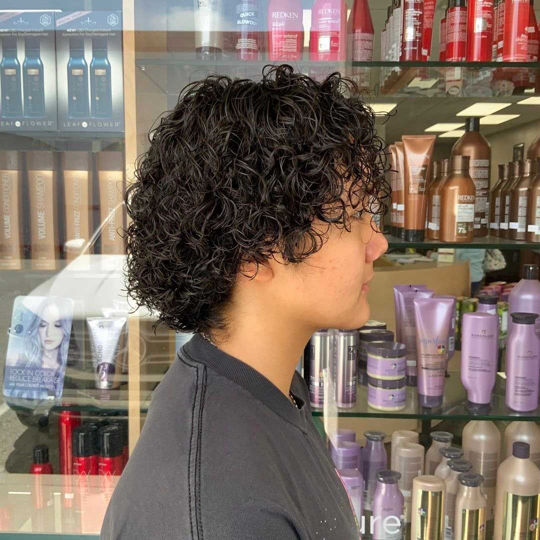 Person with curly dark hair, side profile in a salon. They wear a dark shirt, surrounded by hair products.