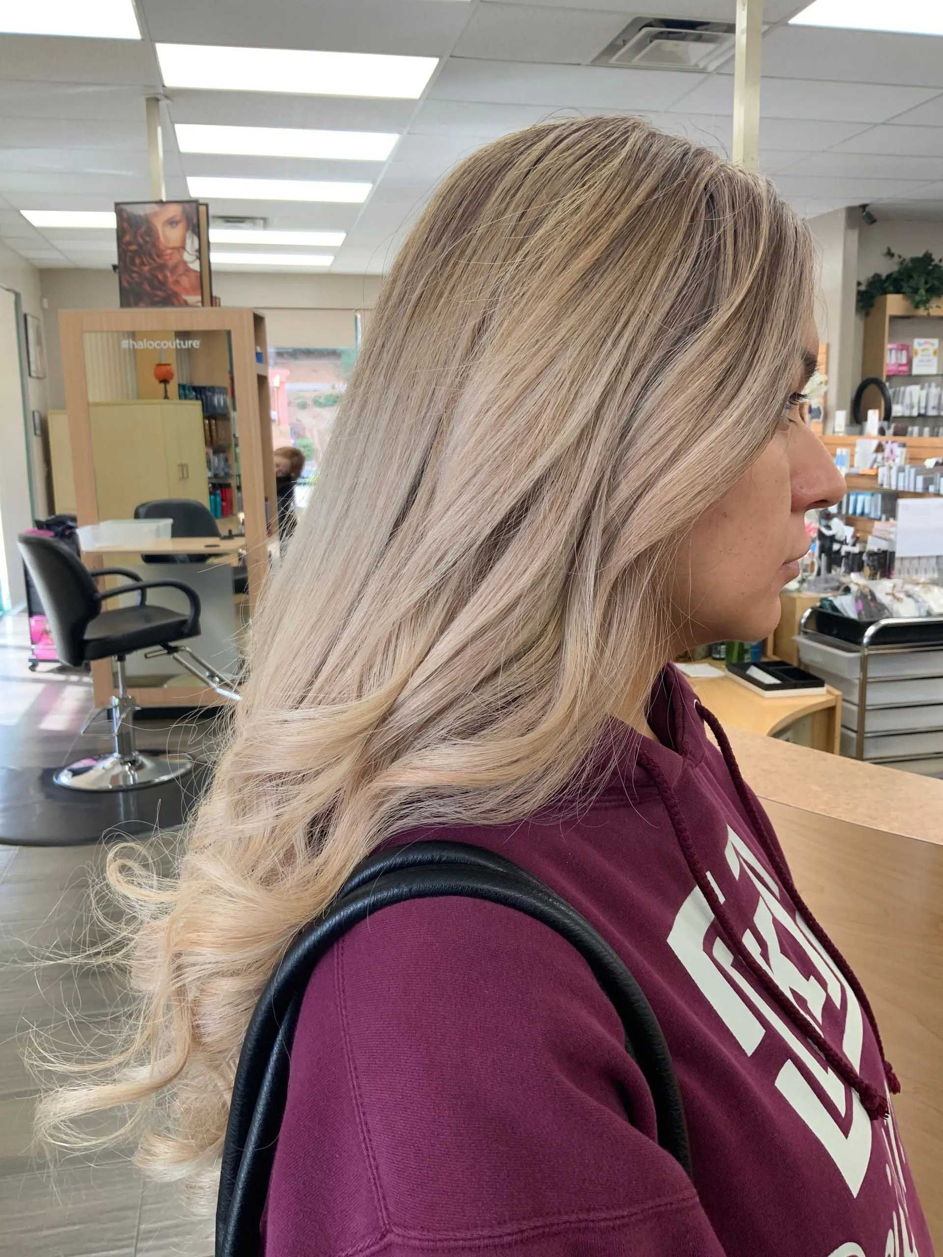 Woman with long blonde hair in a salon, wearing a maroon hoodie and carrying a black purse.