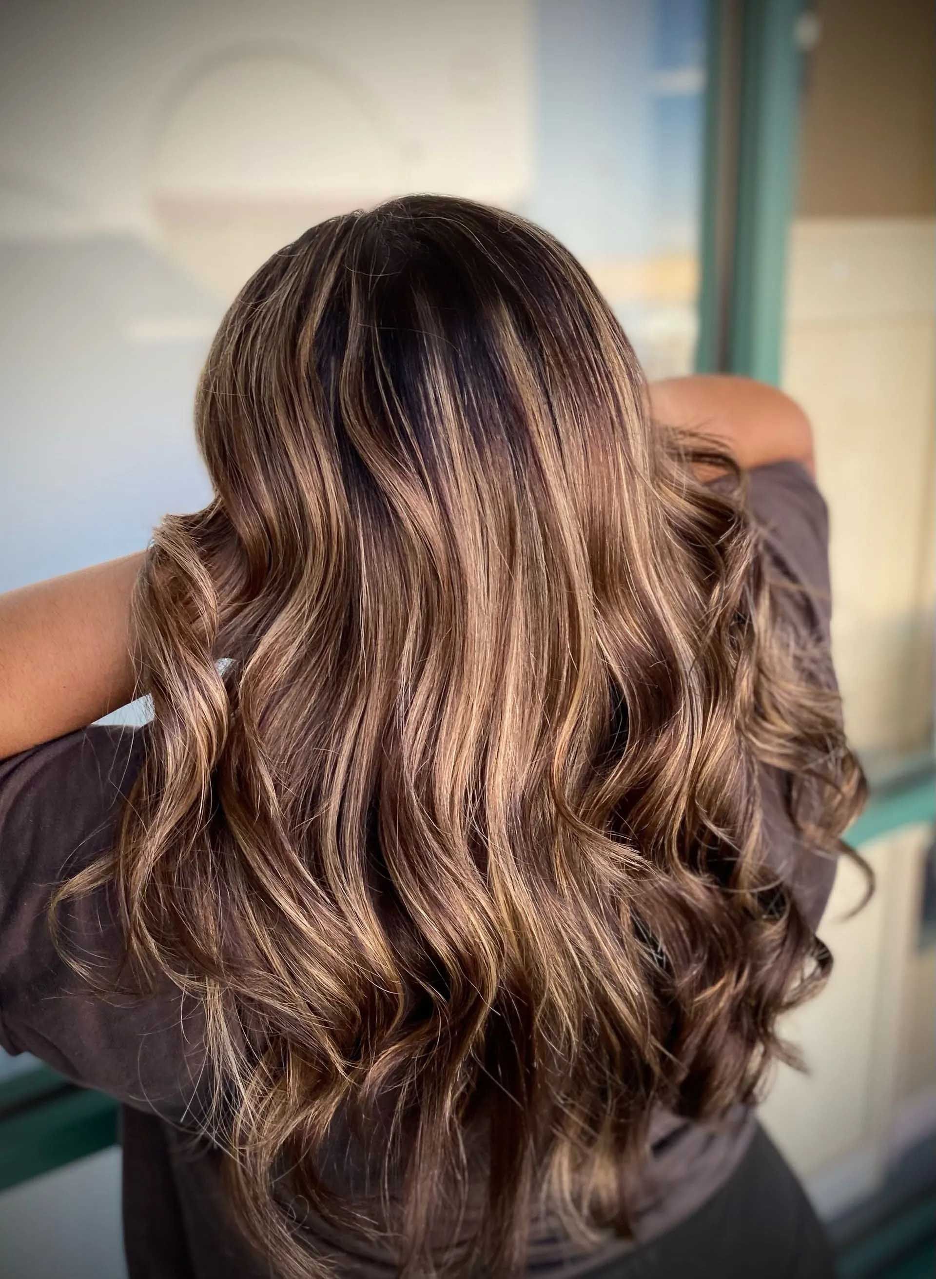 Woman with long wavy brown hair with highlights, arms raised.