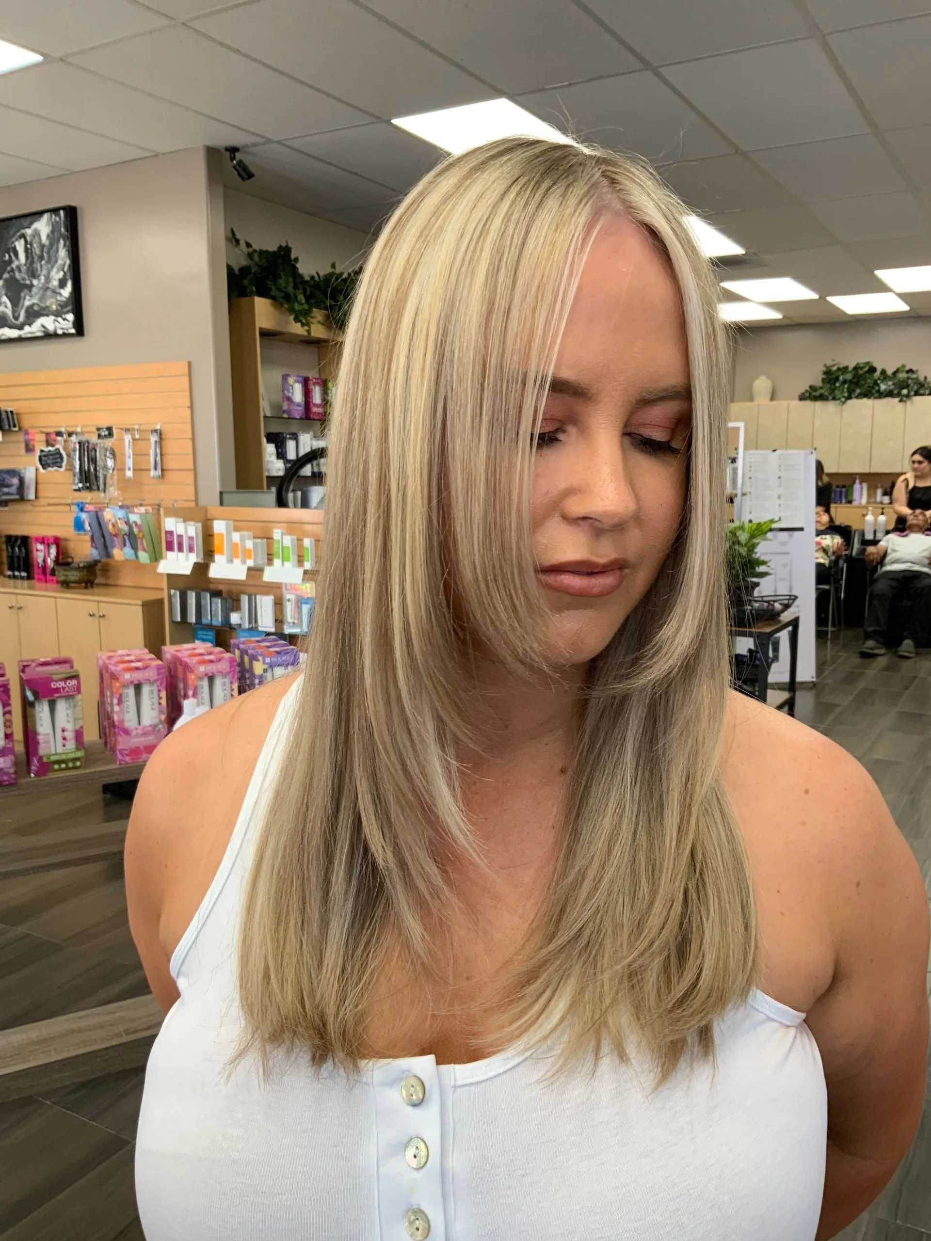 Woman with long blonde layered hair in a salon, wearing a white tank top.