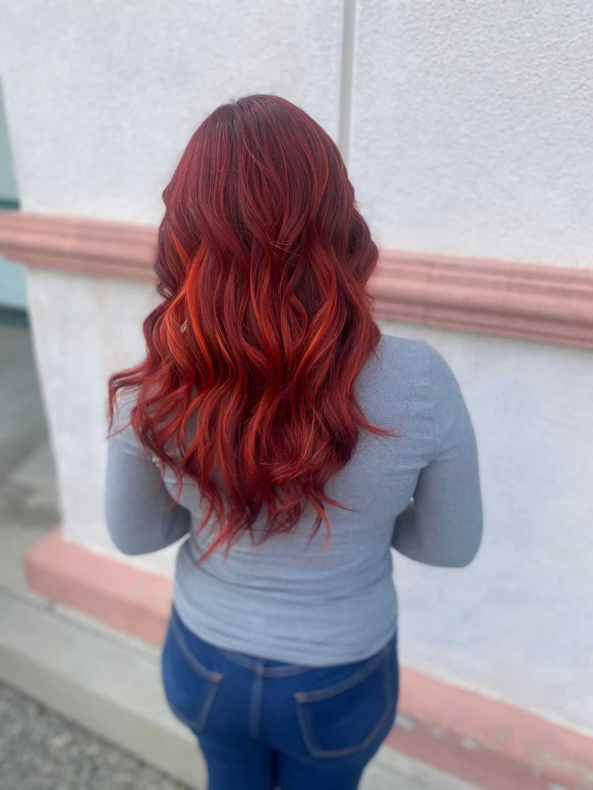 Person with long, red hair styled in waves, wearing a gray long-sleeved shirt and blue jeans, standing outside.