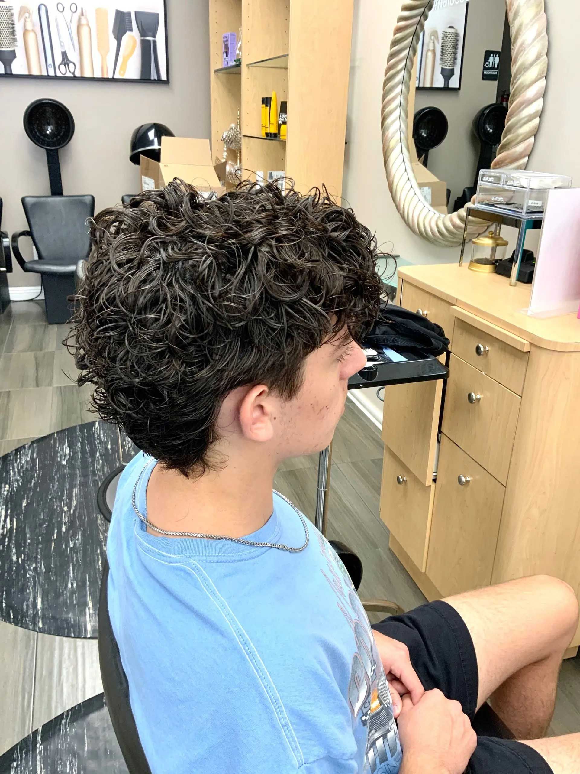 Person with curly hair at a salon, wearing a blue shirt, seated. Interior shot with mirror and styling tools.