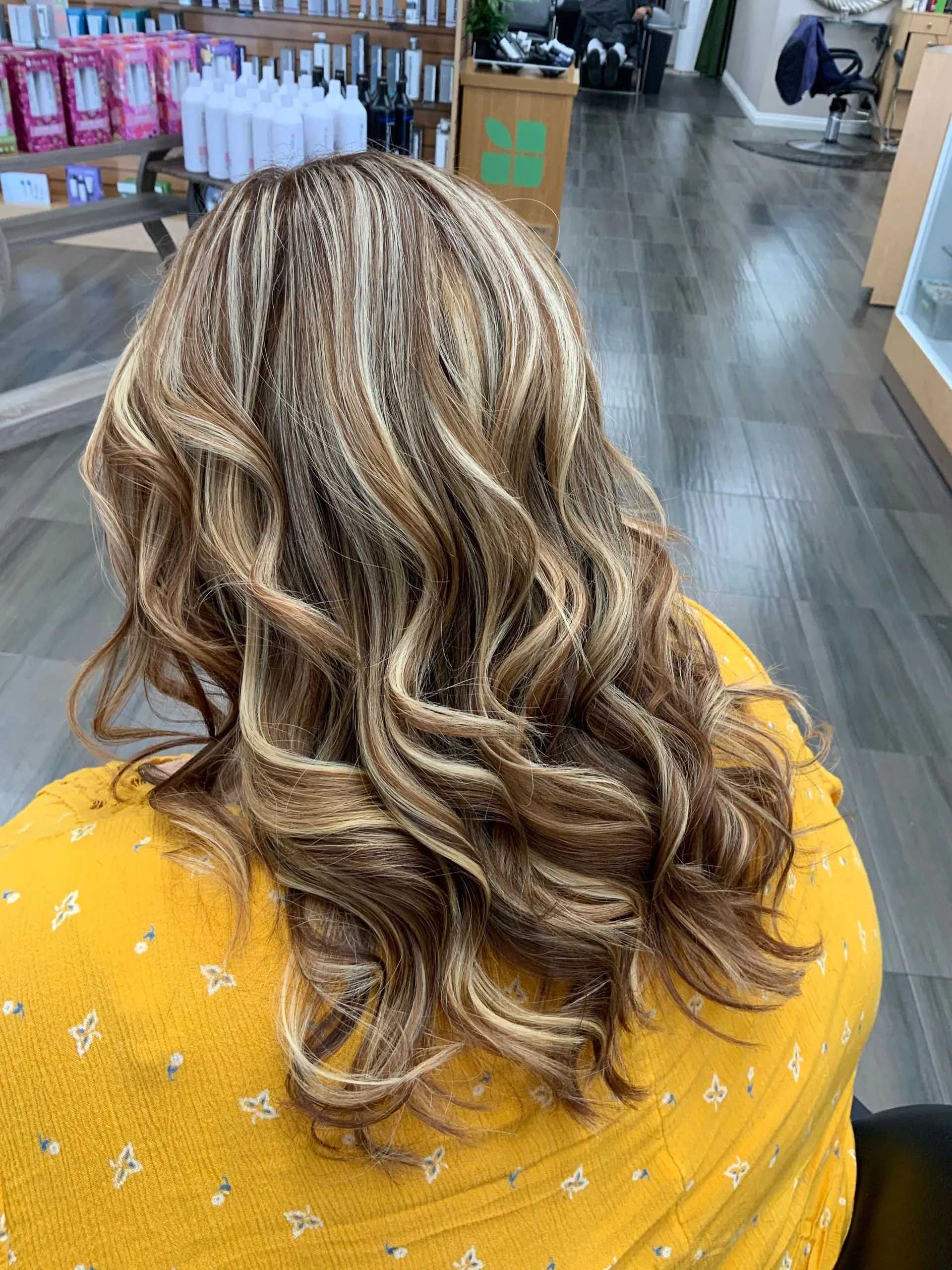 Woman with wavy, highlighted brown hair in a salon, wearing a yellow top.