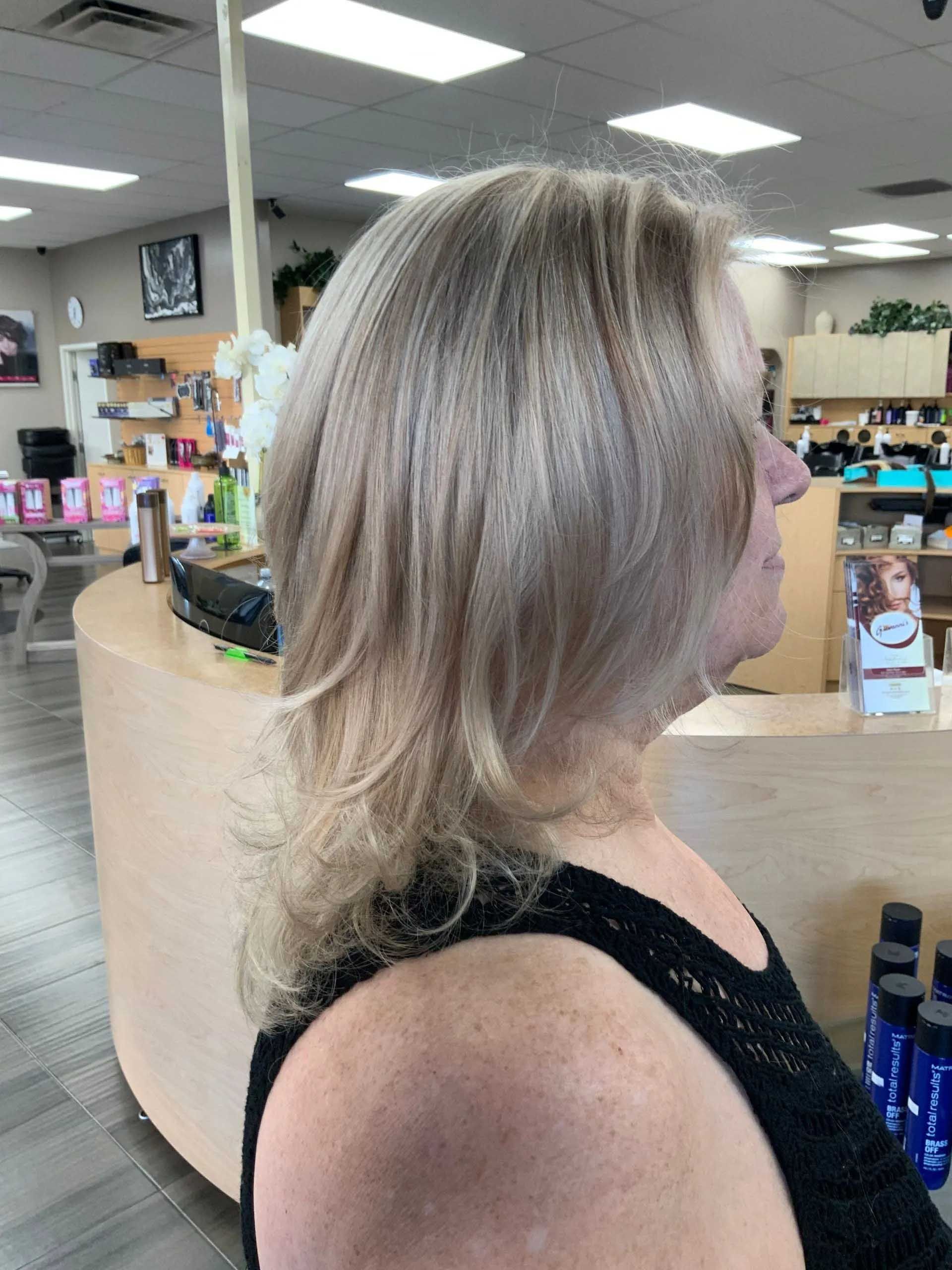 Woman with layered, light blonde hair in a salon, looking right. Wearing a black top.