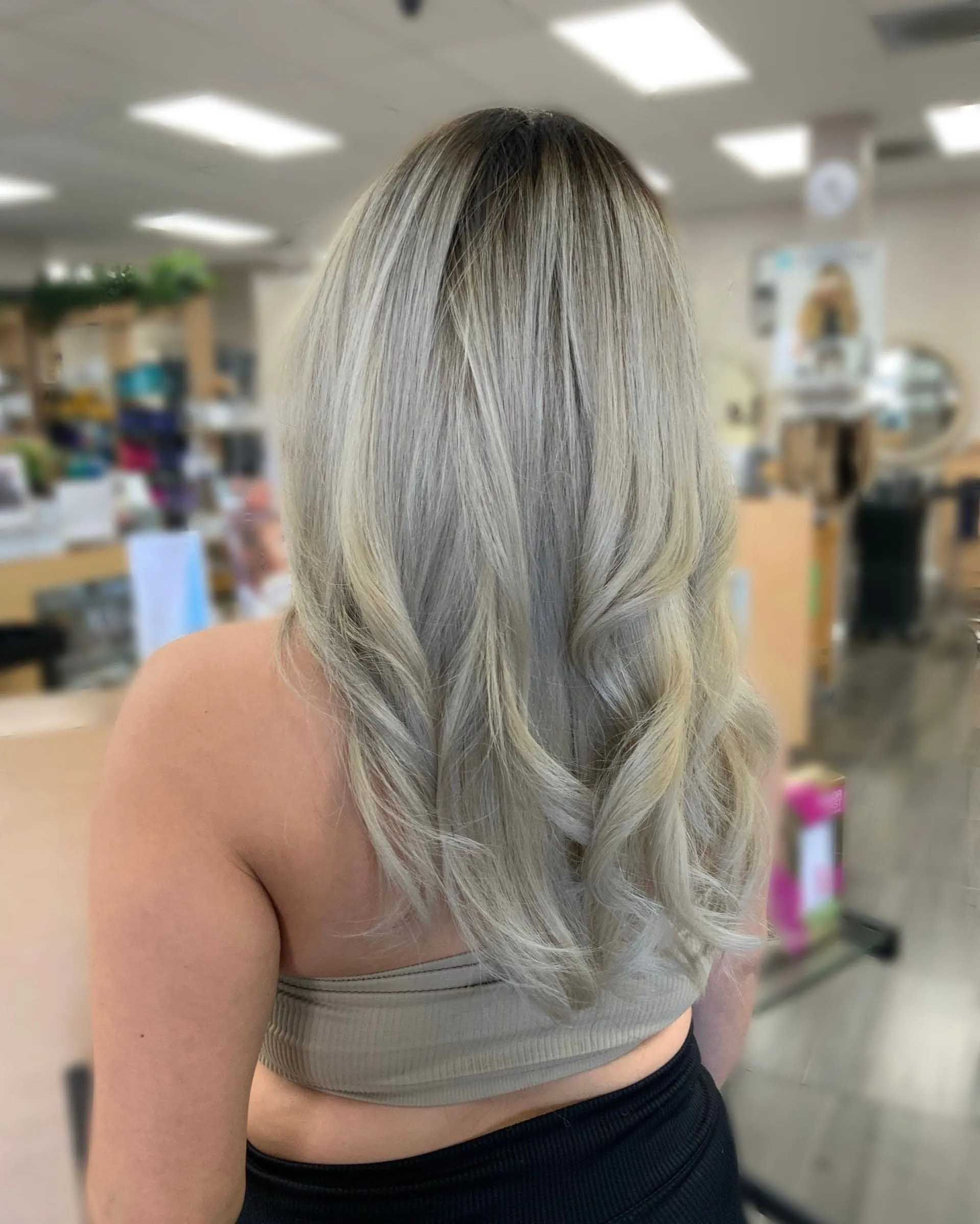 Woman with long, wavy blonde hair; dark roots. She wears a tube top; blurred salon background.