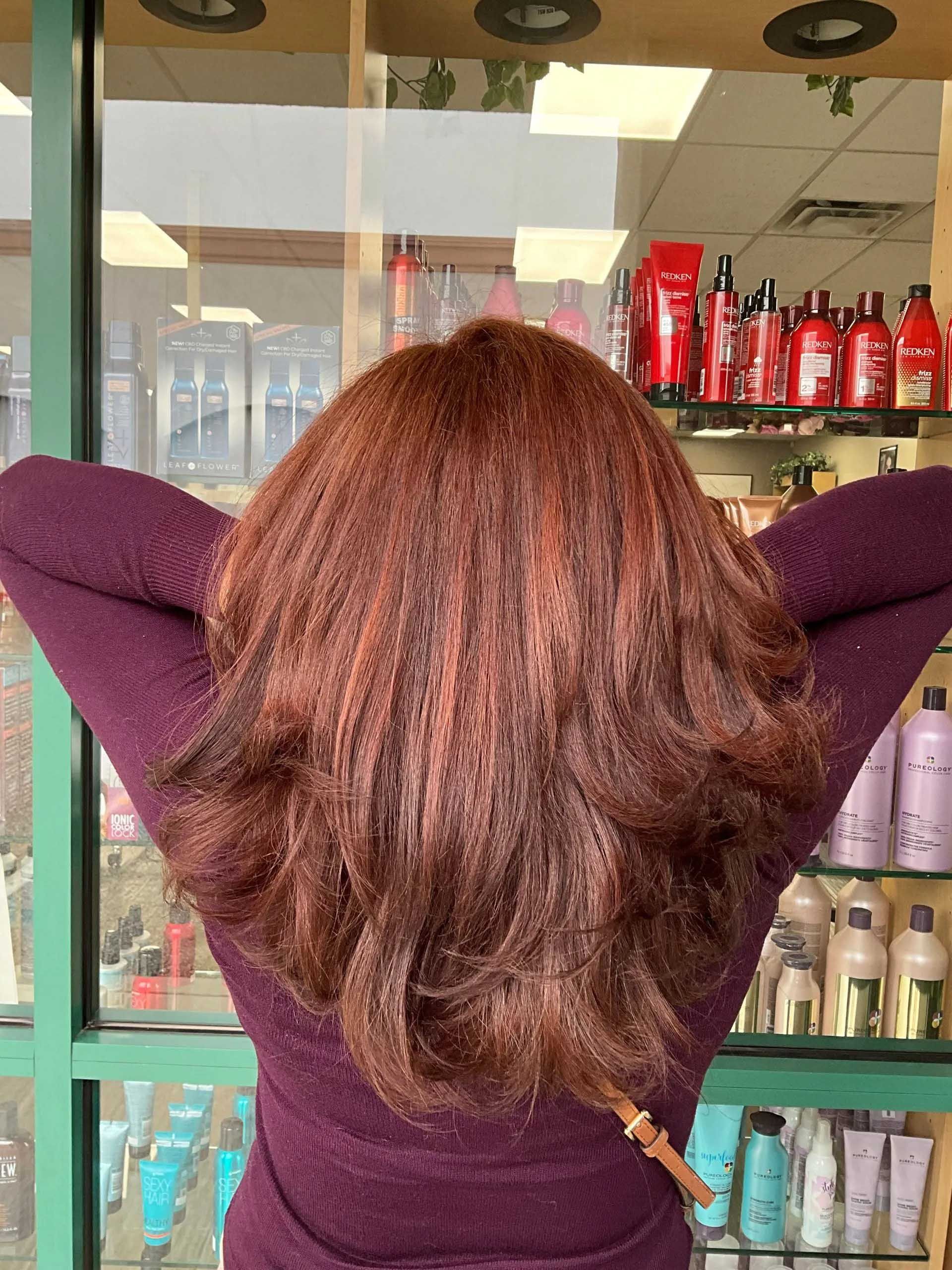 Person with reddish-brown hair, arms raised behind head, in front of hair products.