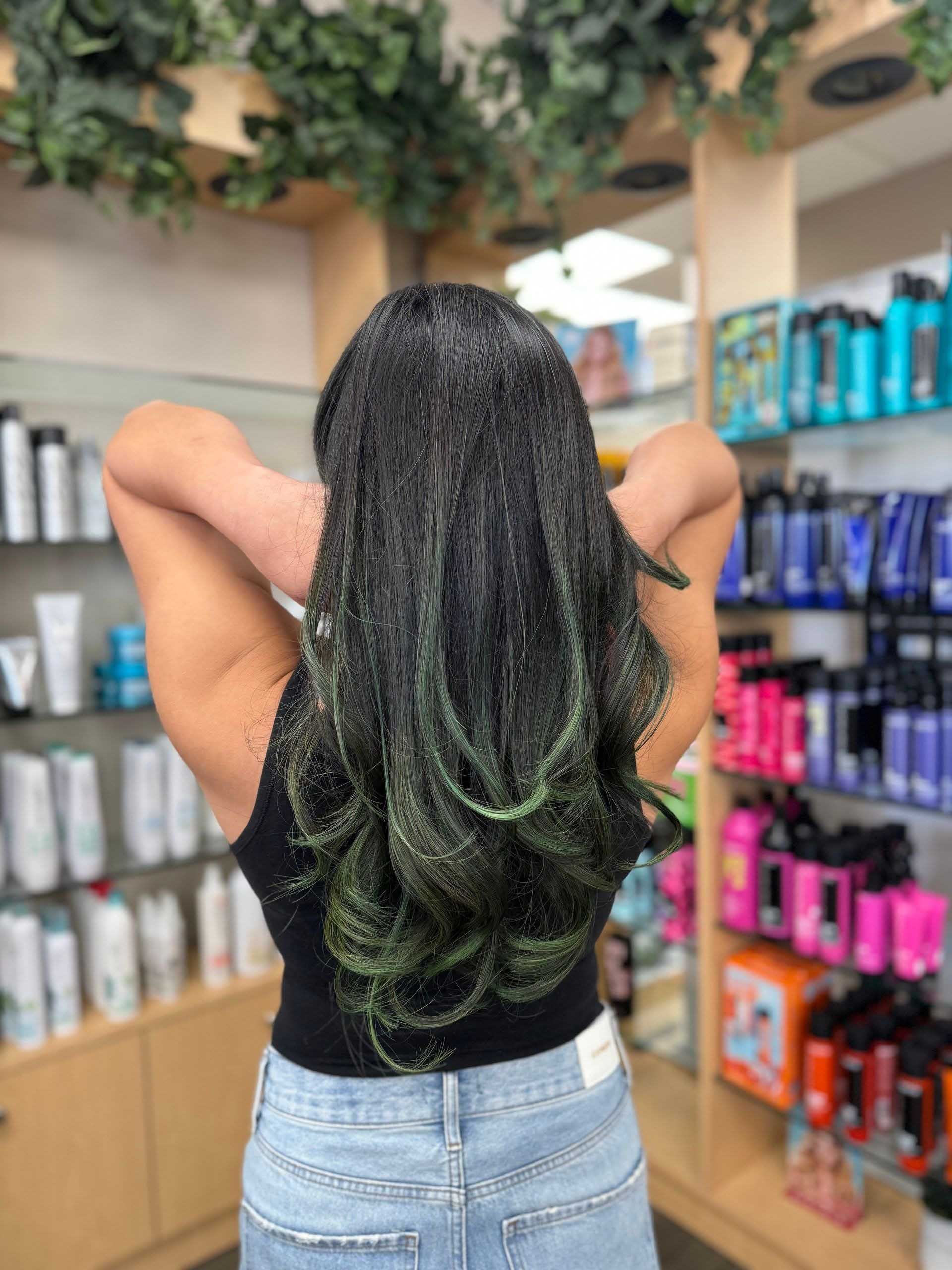 Woman with long black hair, green ombre, hands behind her head, in a hair product store.