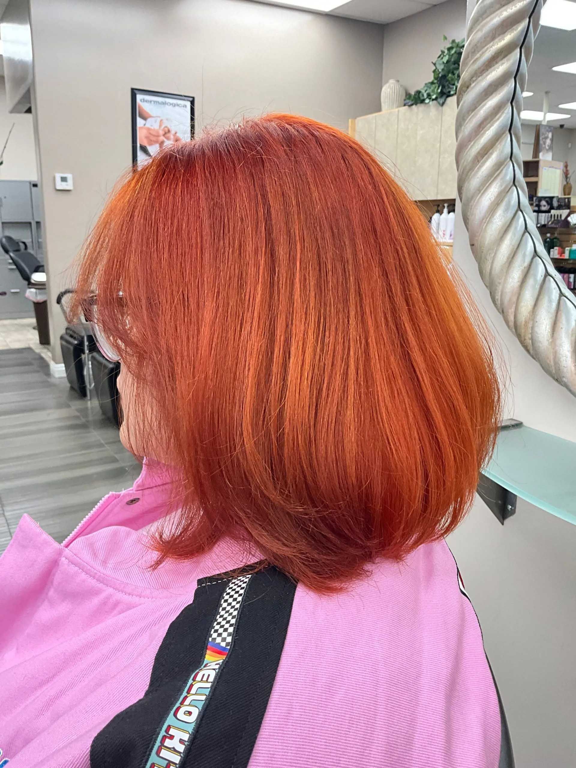 Woman with vibrant orange hair in a salon setting, wearing a pink jacket.