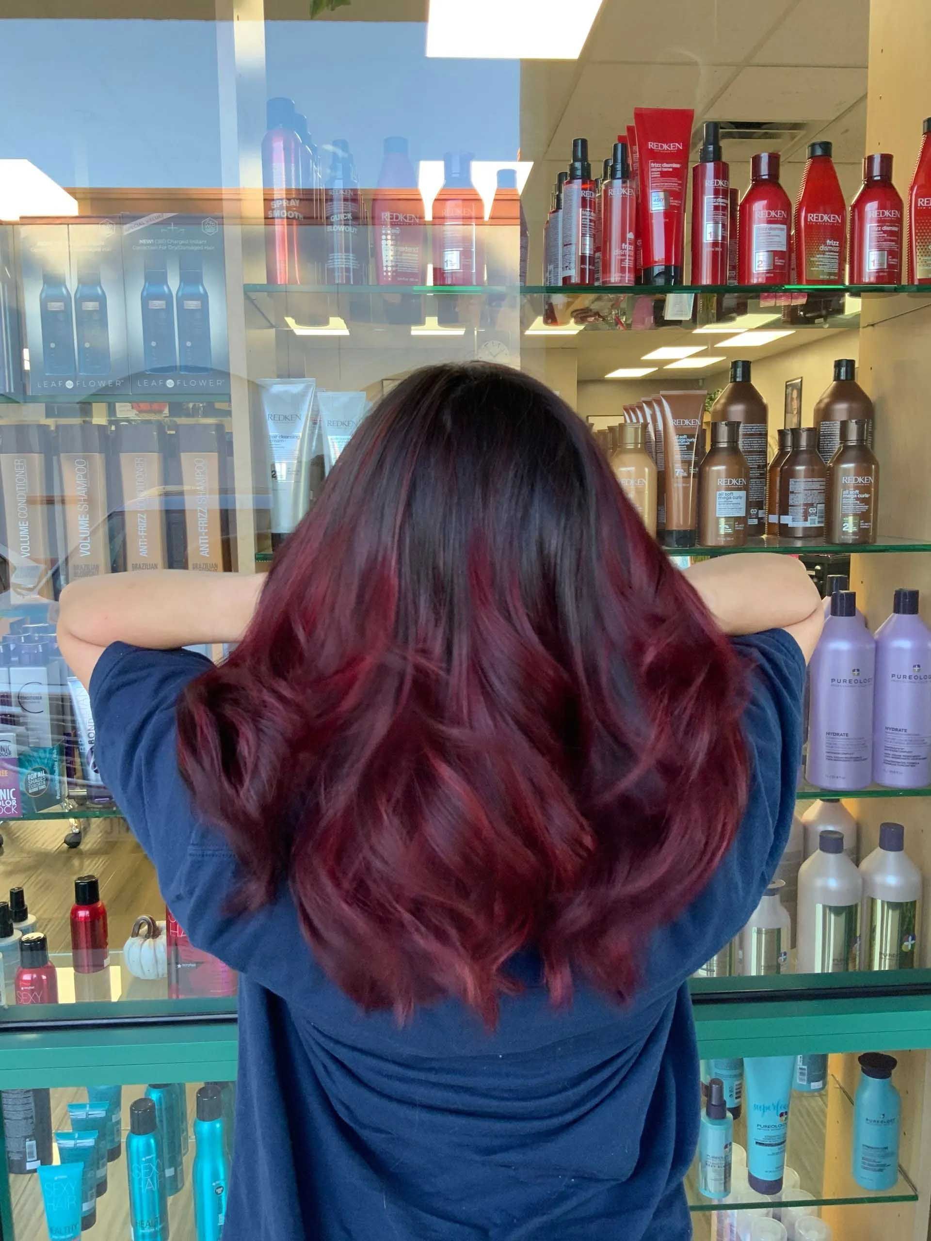 Person with dark hair dyed red, arms crossed, in front of hair products in a salon.