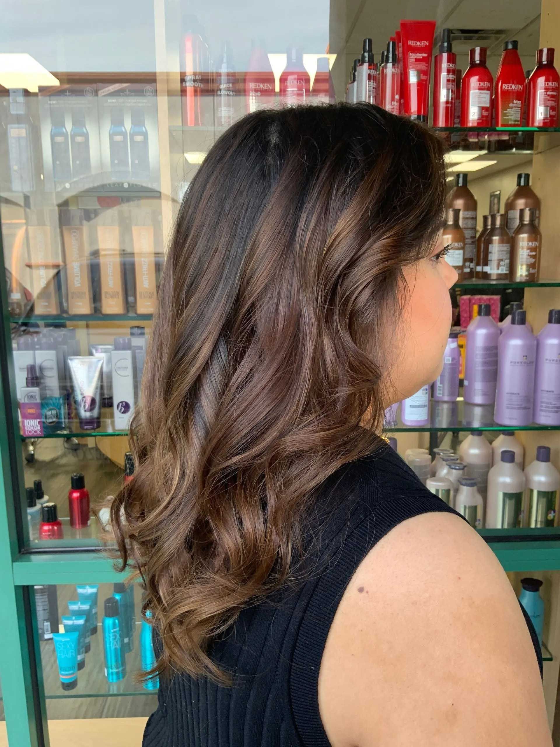 Woman with wavy, dark brown hair in a salon, facing away from the camera, near shelves of hair products.