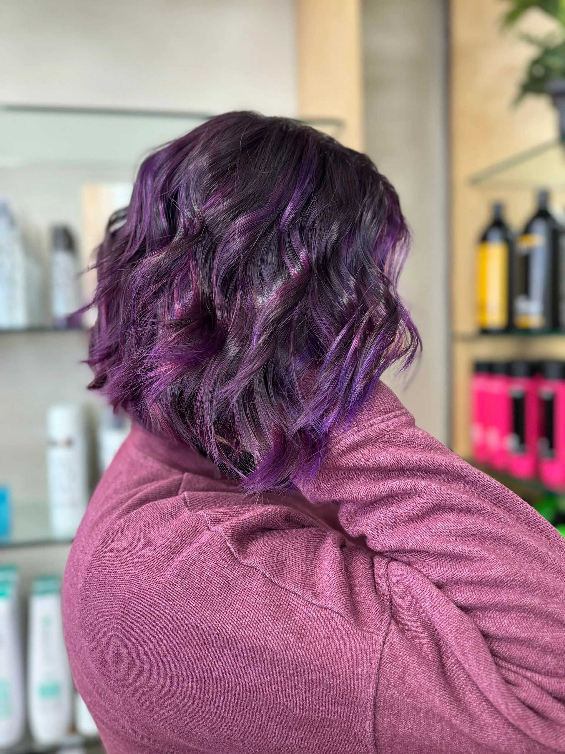 Woman with wavy, purple-streaked hair, wearing a purple sweater, in a salon.