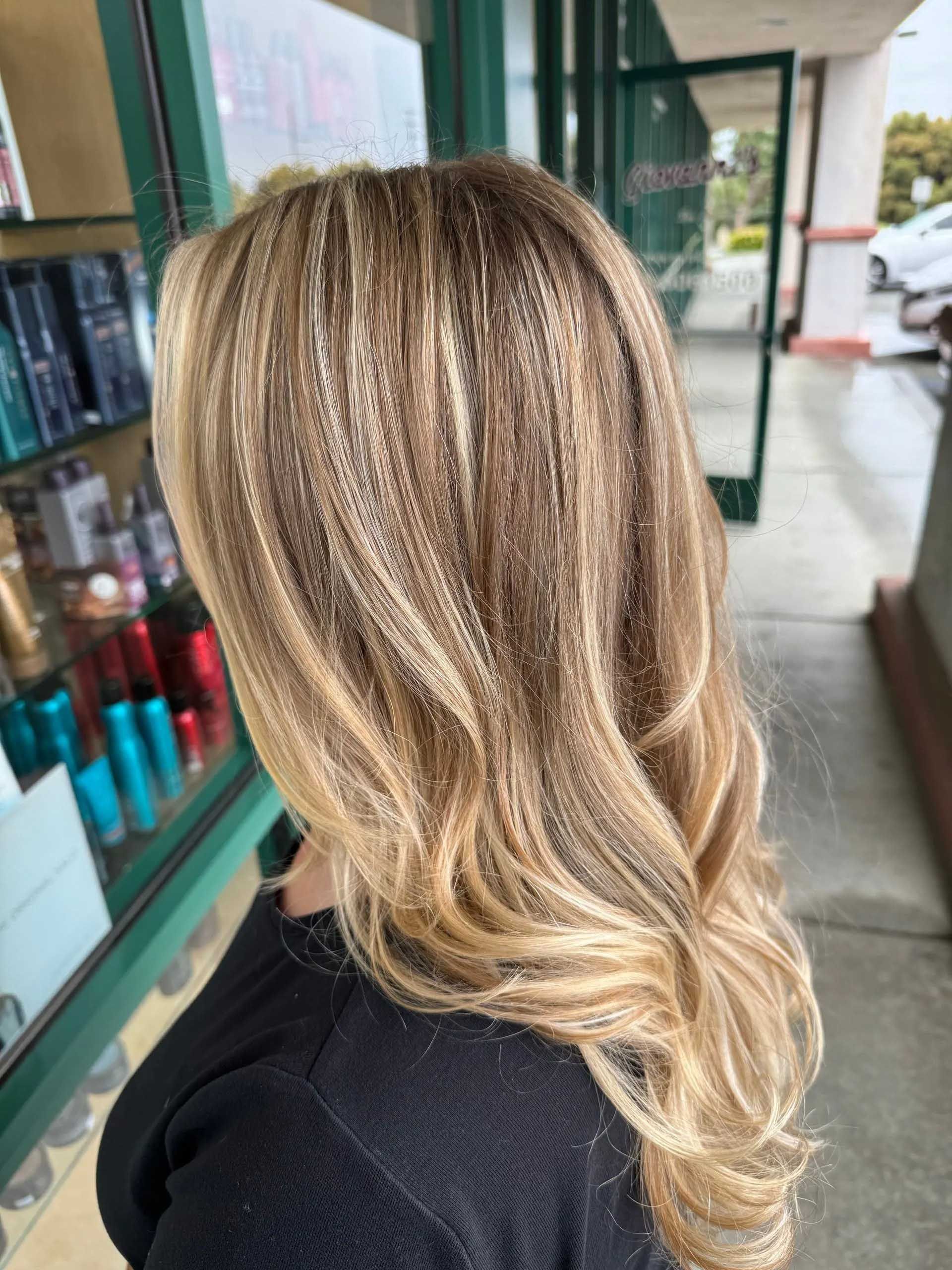 Woman with long, wavy blonde hair with highlights, standing outside a shop with green trim.