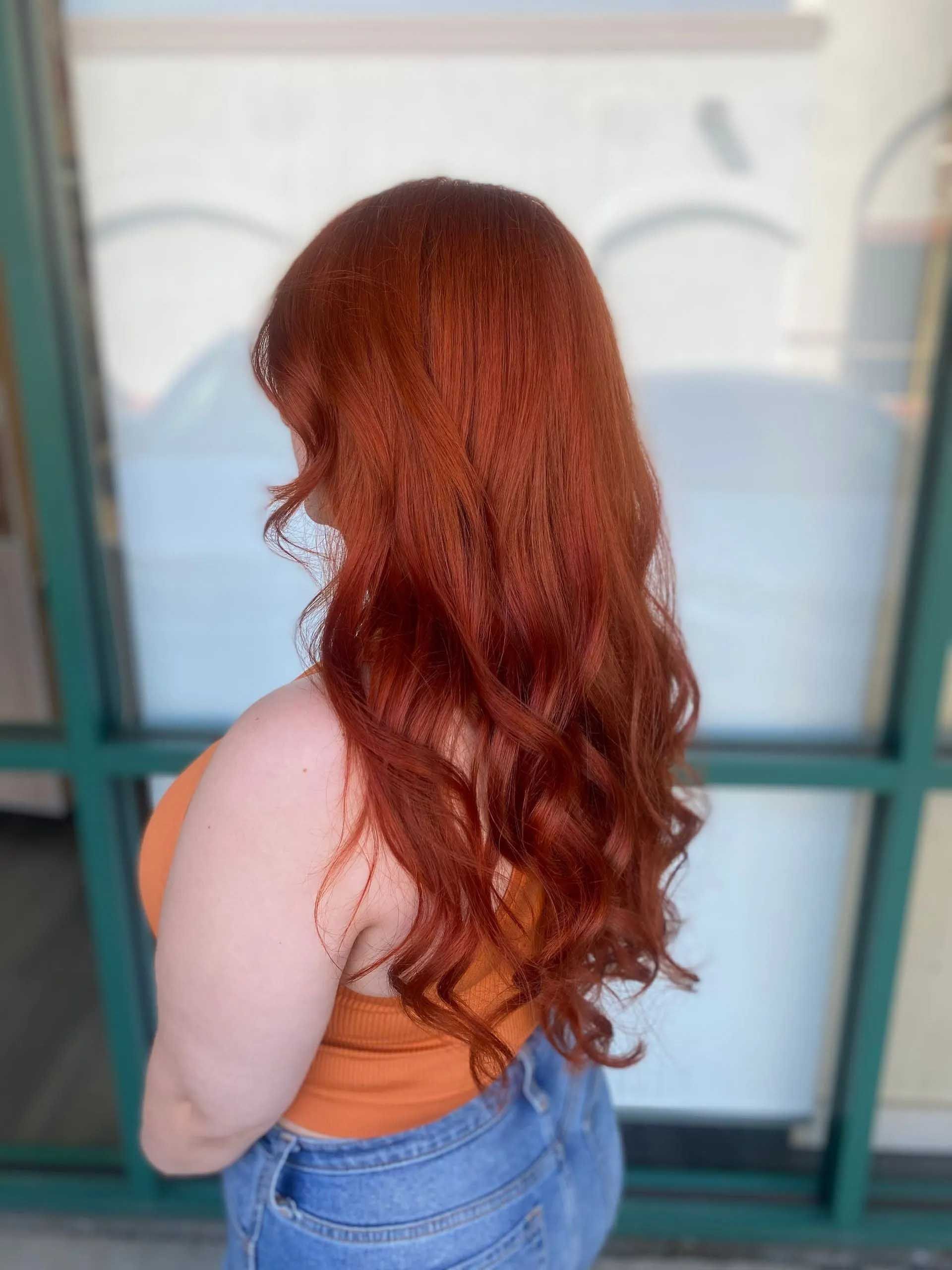 Woman with long, wavy red hair, wearing an orange top and jeans, stands in front of a window.