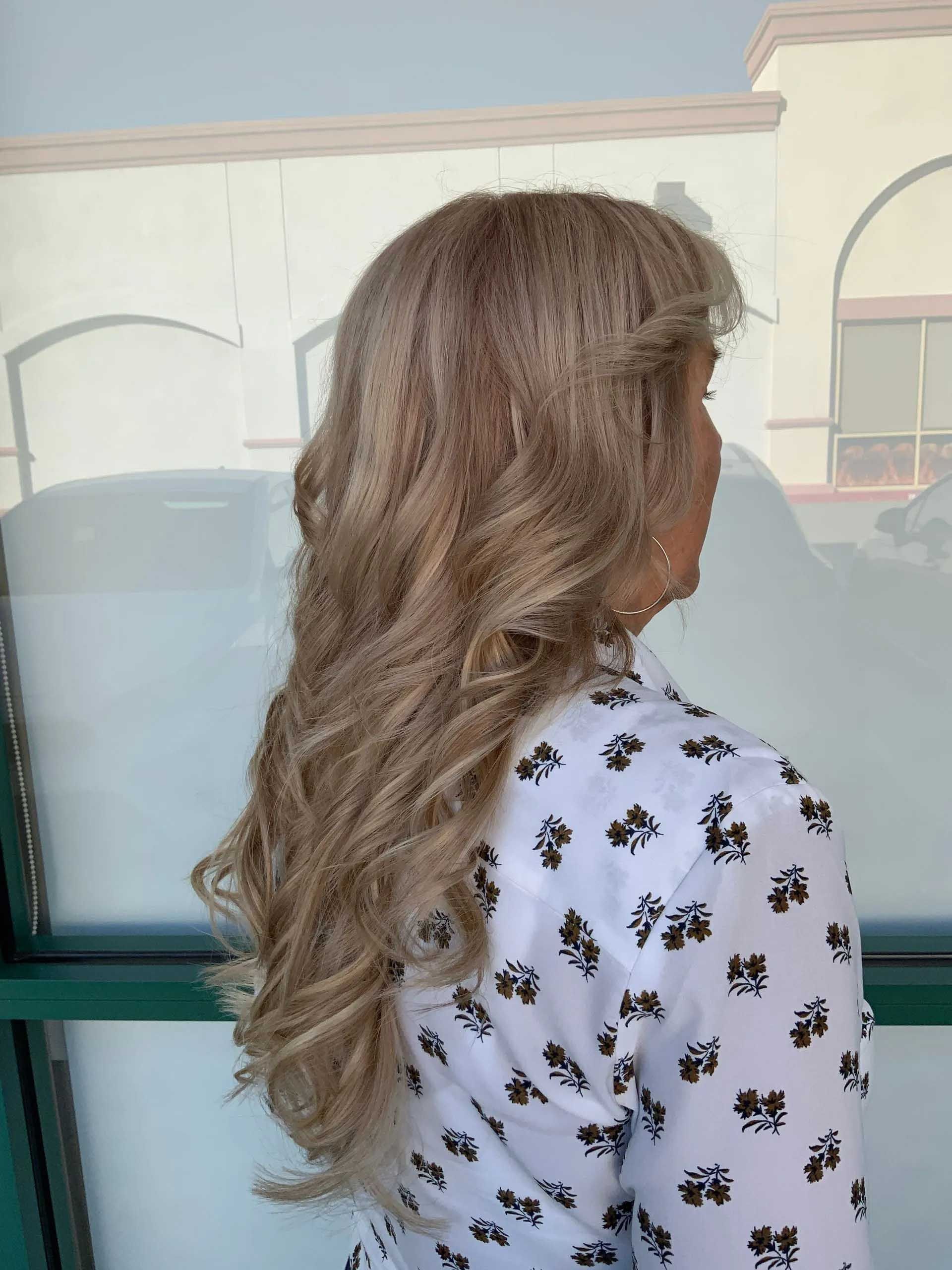 Woman with long, wavy blonde hair and a white patterned blouse, partially facing away.