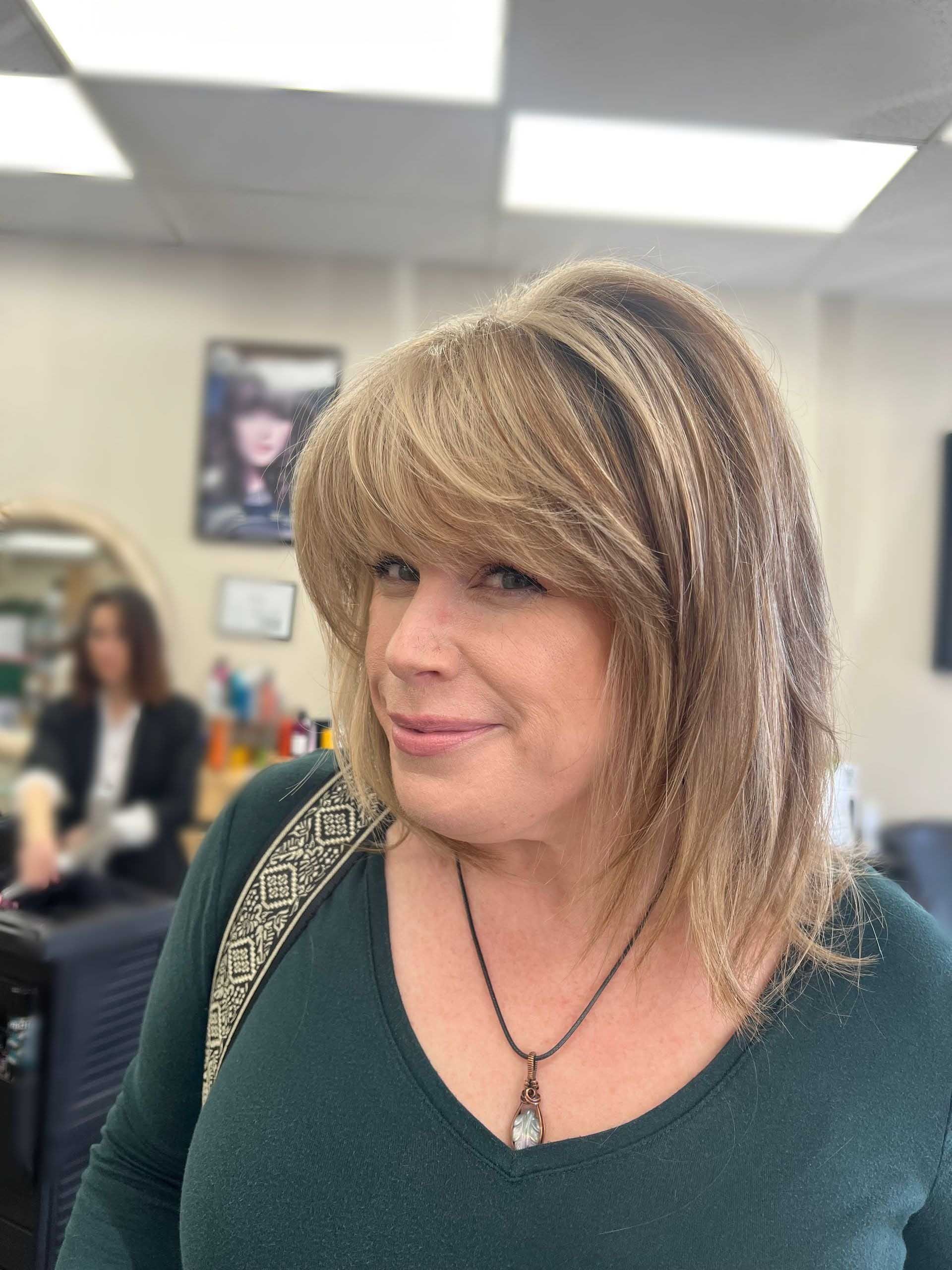 Woman with blonde layered hair and bangs in a salon, wearing a green sweater and necklace, smiling.