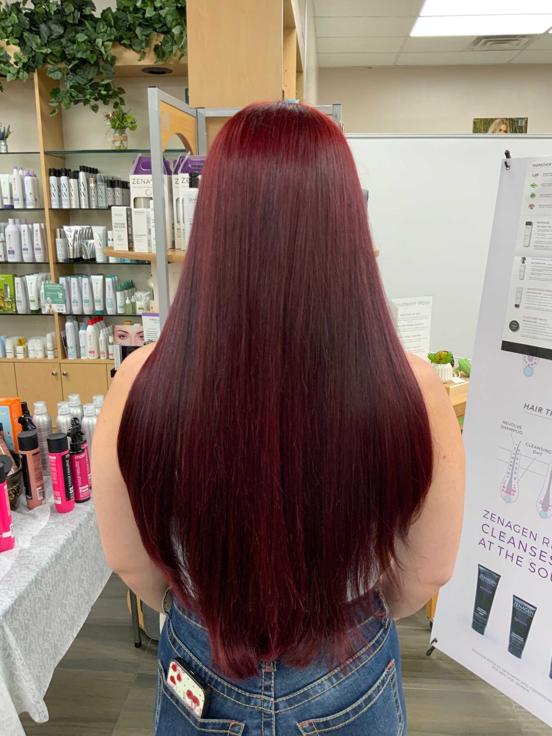 Woman with long, straight, burgundy-colored hair. Standing in a salon with products on shelves behind her.