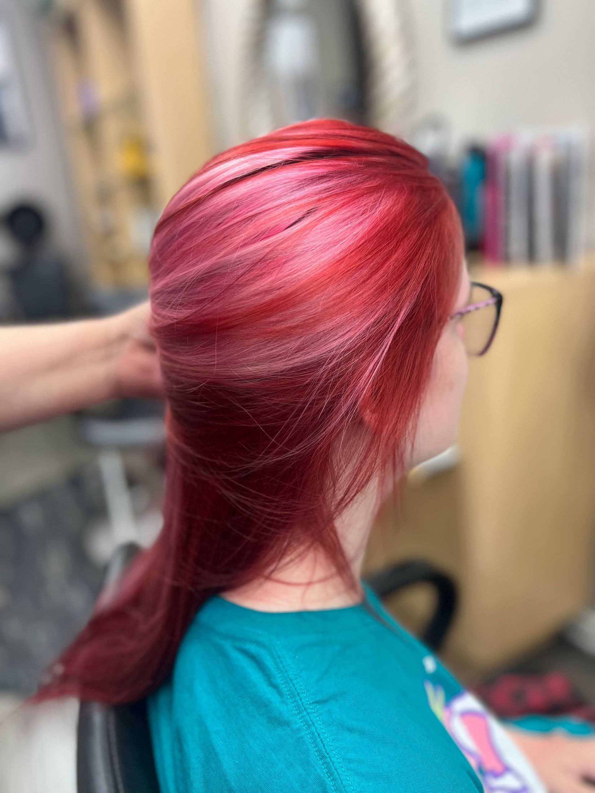Woman with long, vibrant red hair sits in a salon chair. A stylist's hand is visible near the hair.