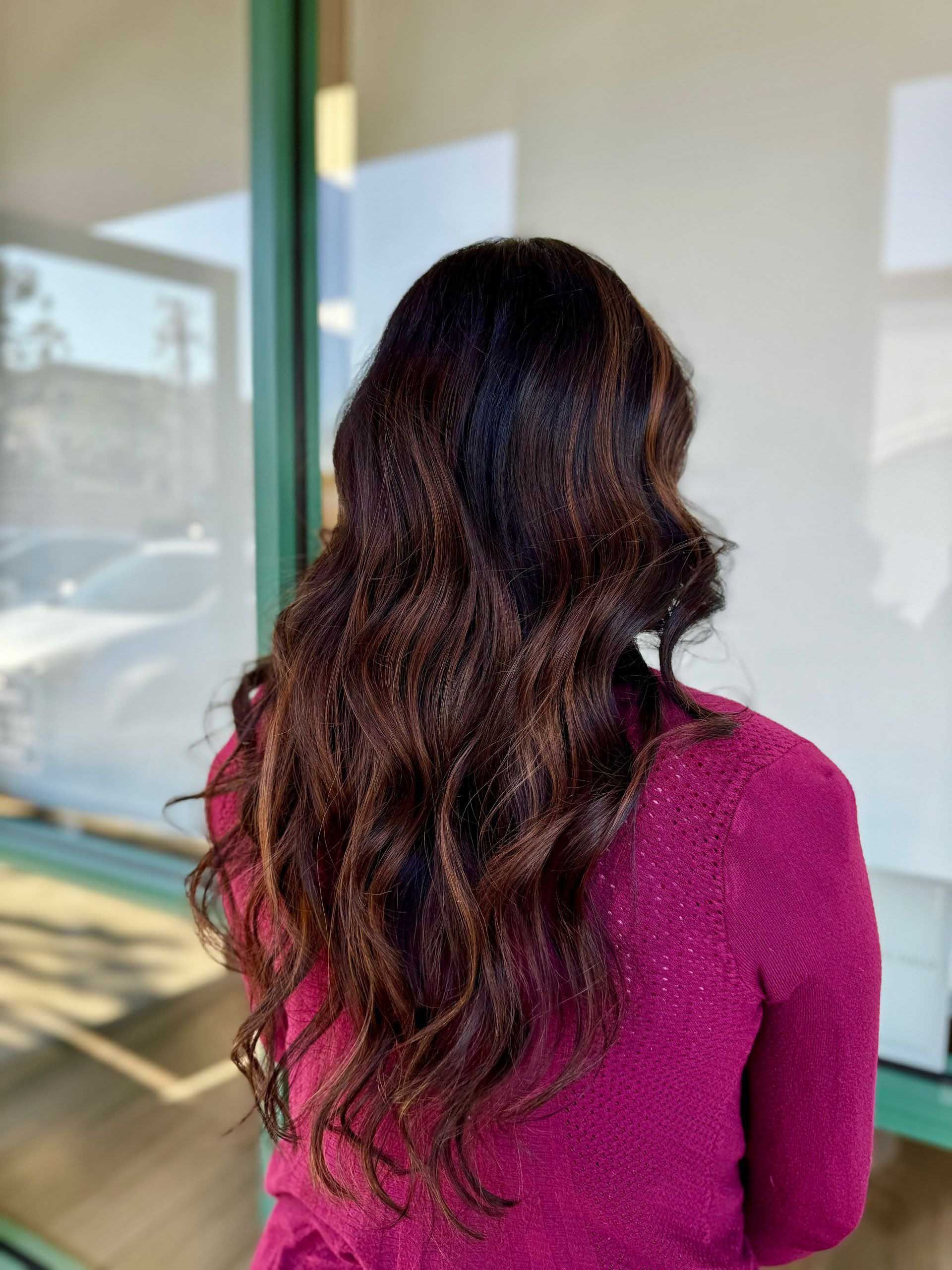Woman with long, wavy brown hair with highlights, wearing a burgundy sweater, standing near a window.