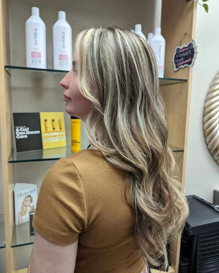 Woman with blonde highlights and wavy hair in a salon, facing left.