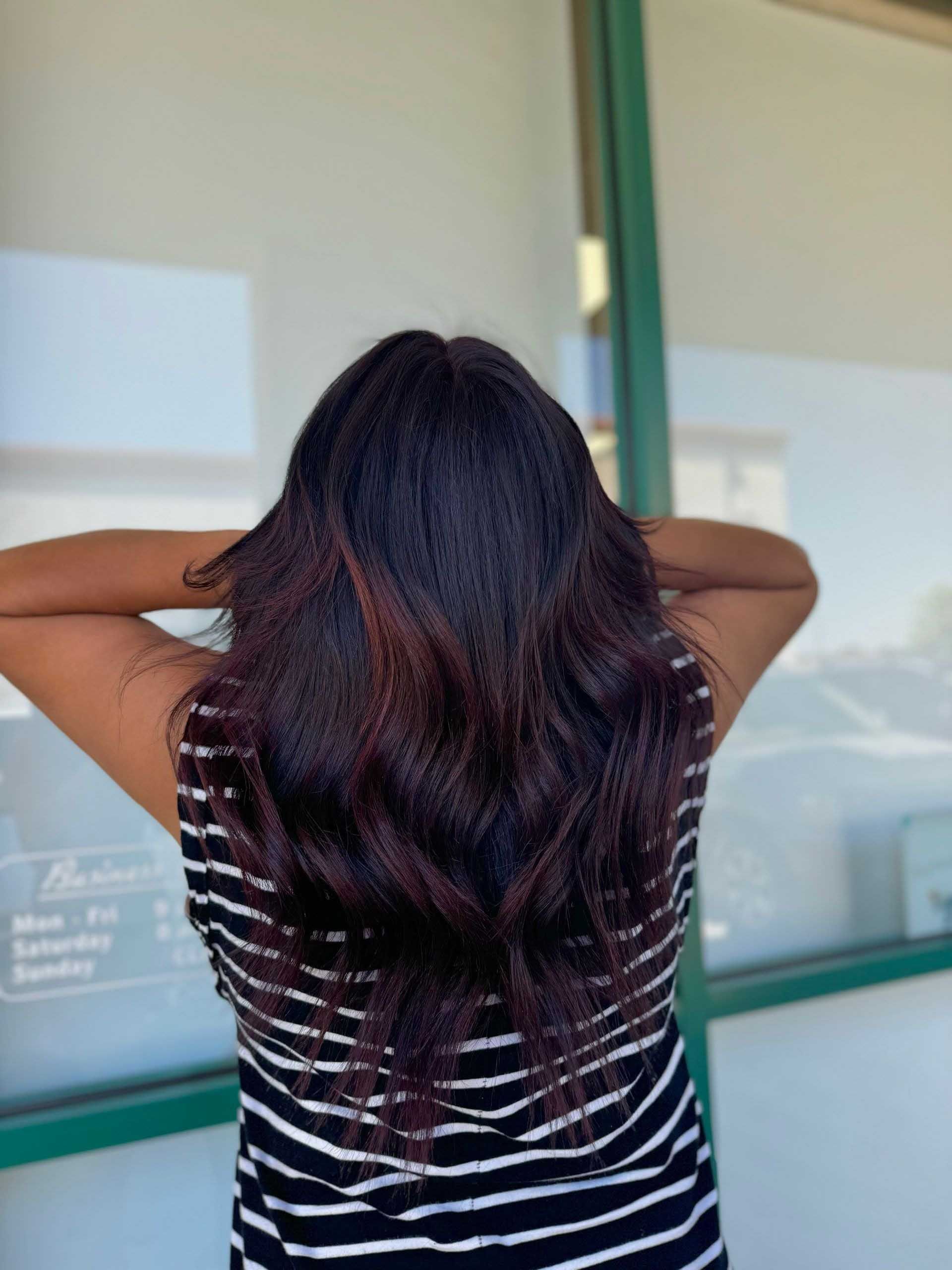 Woman with long dark hair, hands behind head, wearing a striped top, standing outdoors.