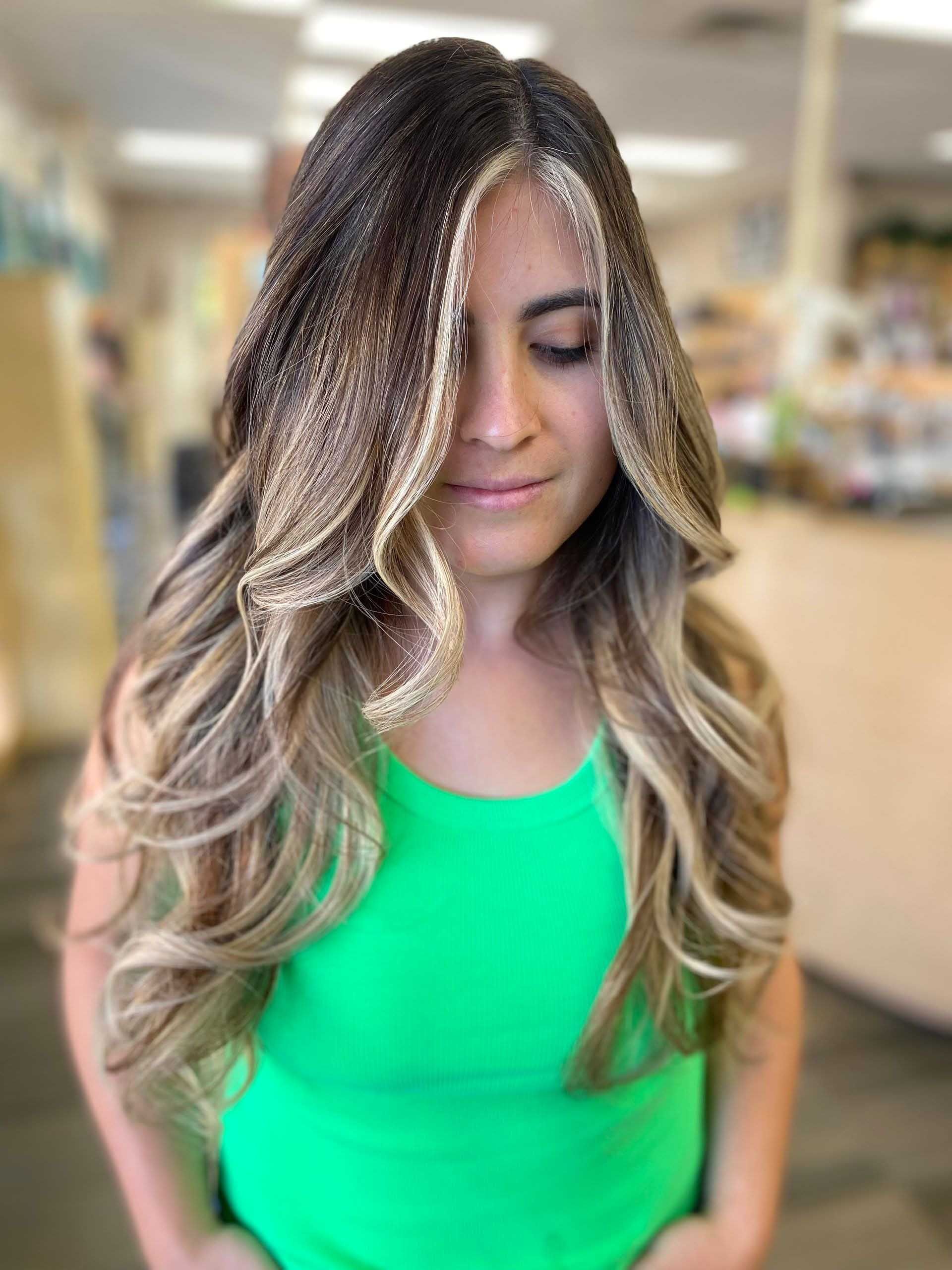 Woman with long, wavy, two-toned hair and a green tank top, eyes closed, in a store setting.