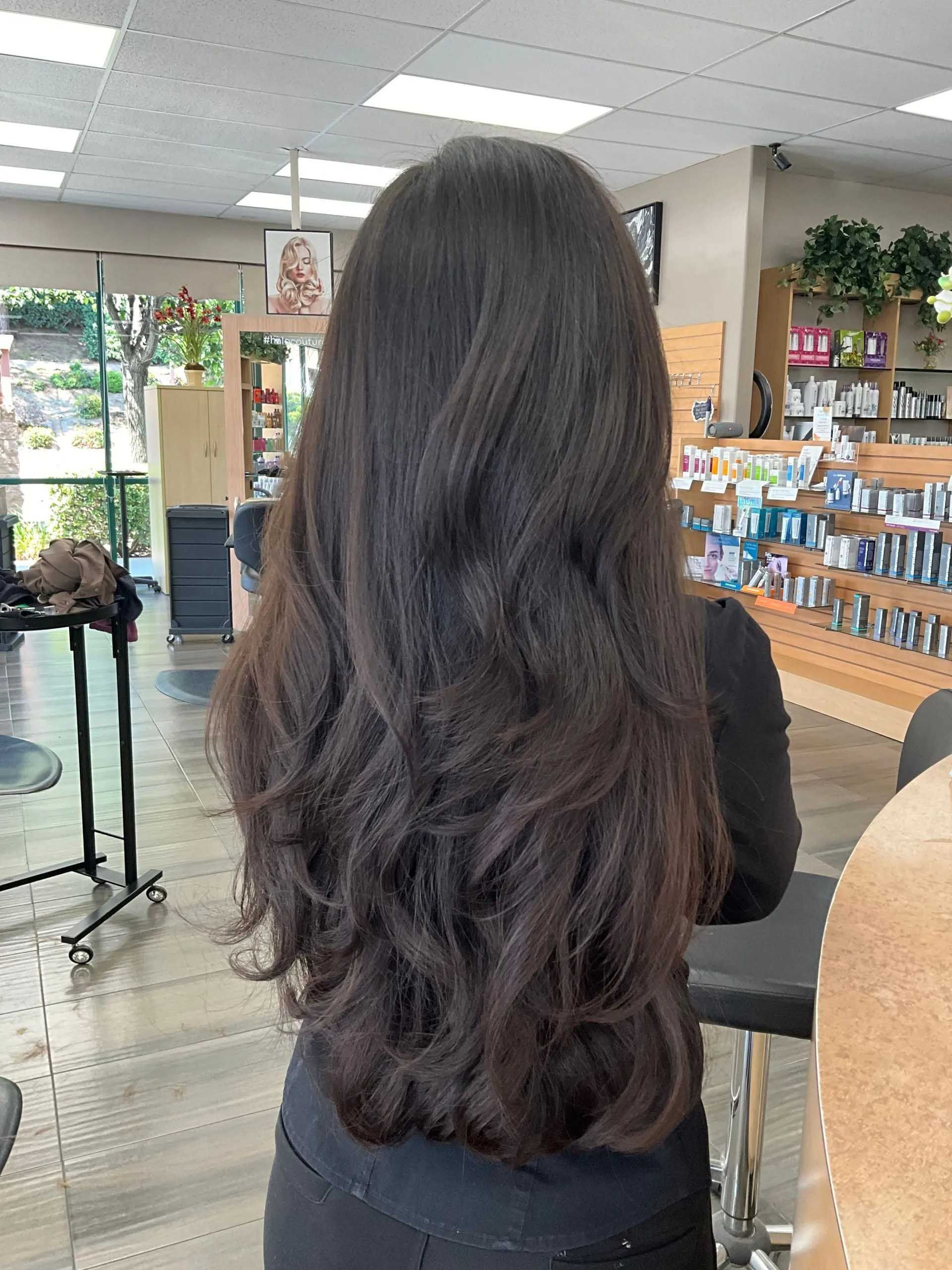 Woman with long, dark layered hair in a salon; standing, back to the camera.