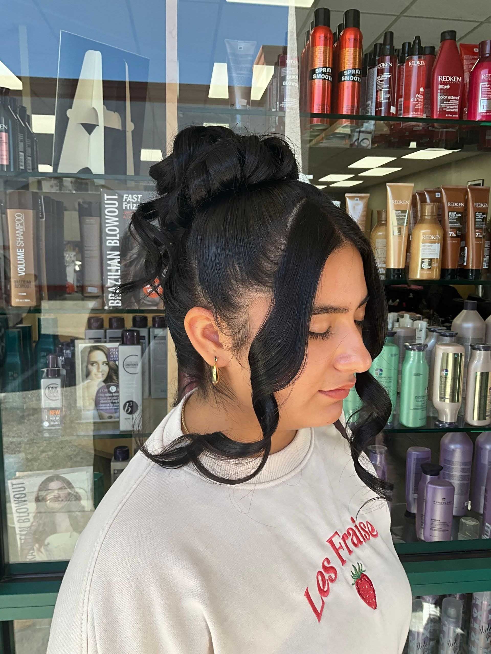 Woman with a dark updo hairstyle, bangs, and wearing a light-colored top, posed inside a hair salon.
