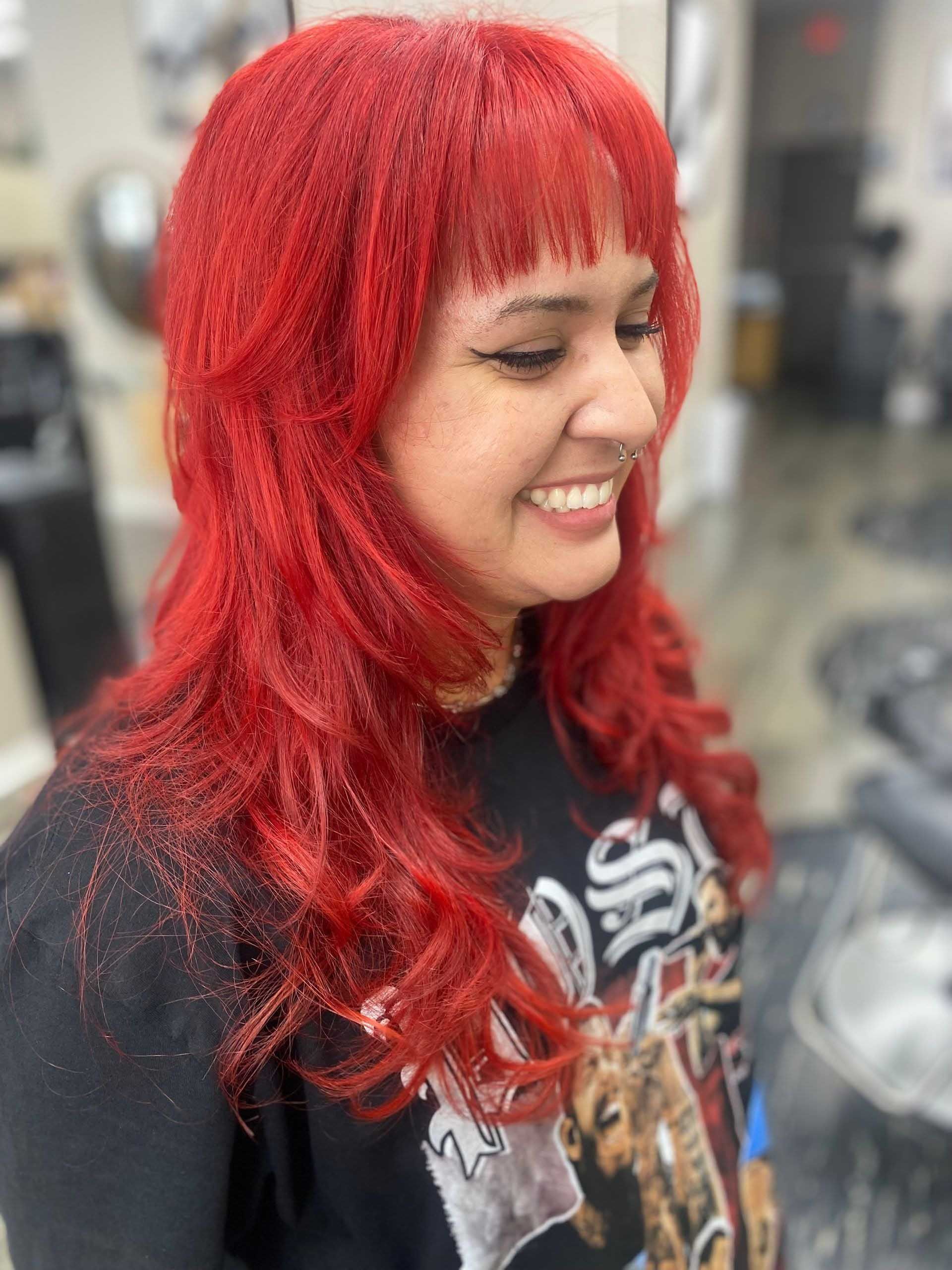 Woman with bright red hair and bangs smiling. Wearing a black graphic tee. Salon setting.
