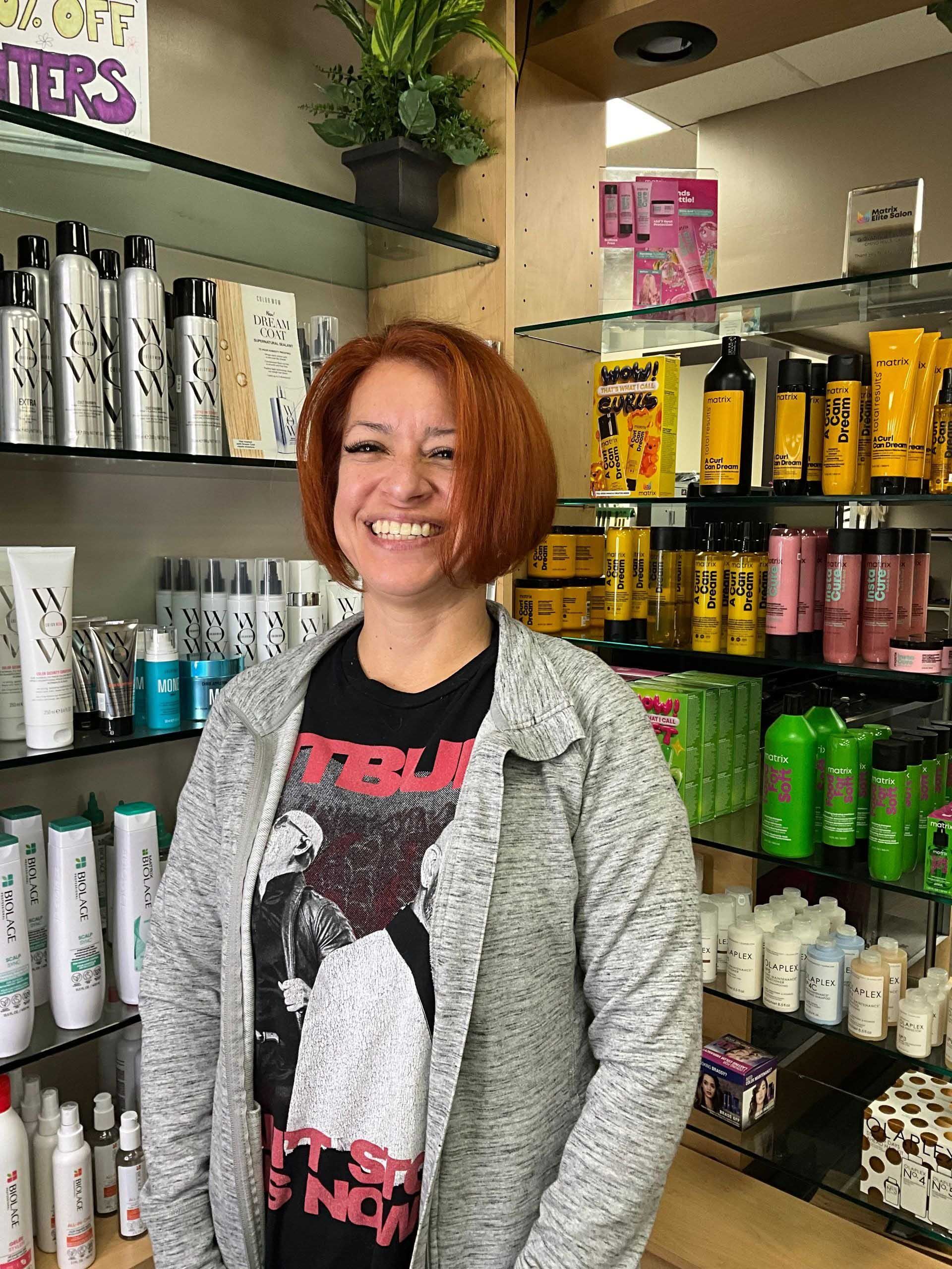 Woman smiling, standing in a shop filled with hair products on shelves. She wears a grey jacket and t-shirt with print.