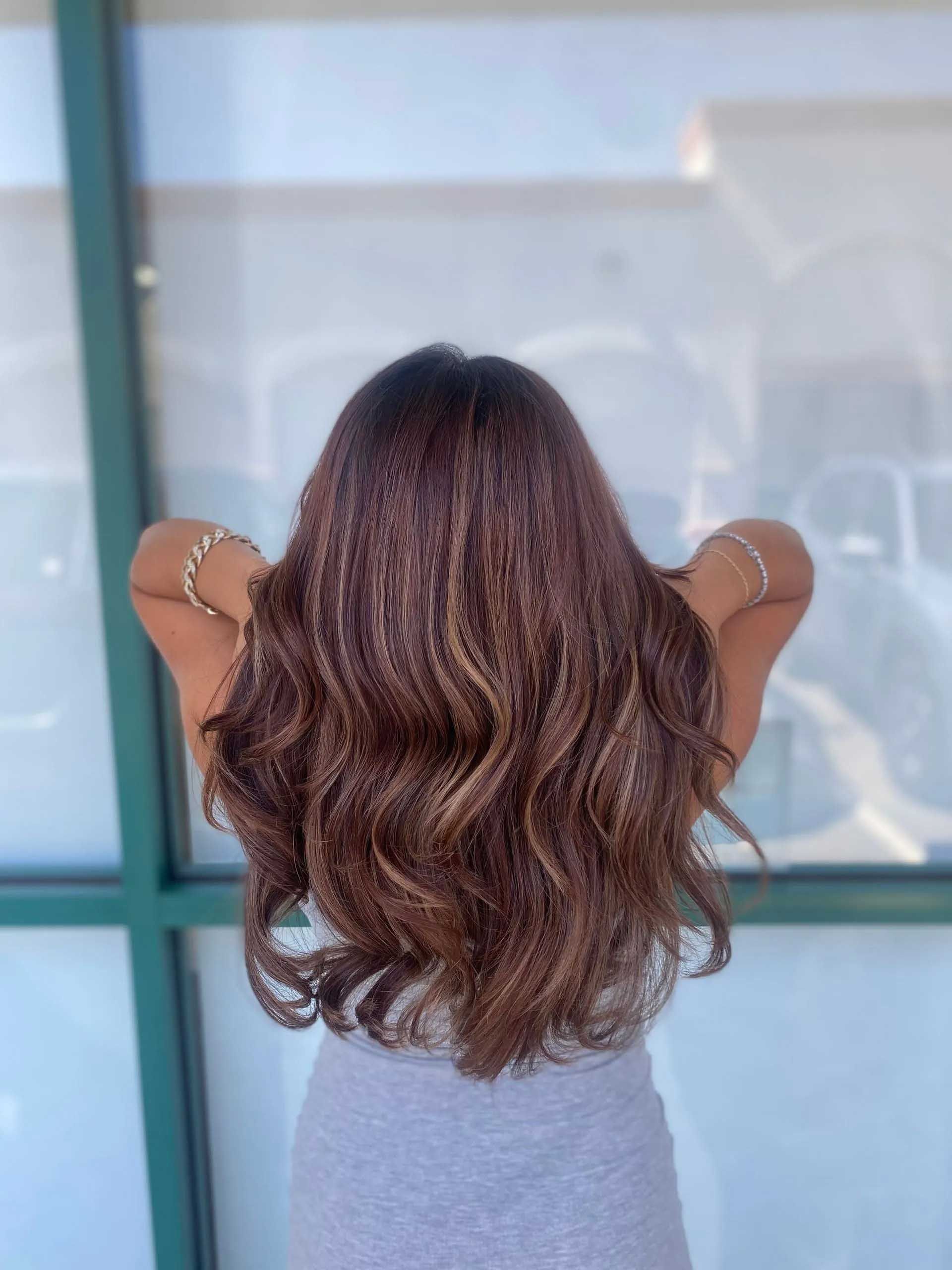 Woman with long, wavy brown hair with highlights, arms raised. Standing in front of a window.
