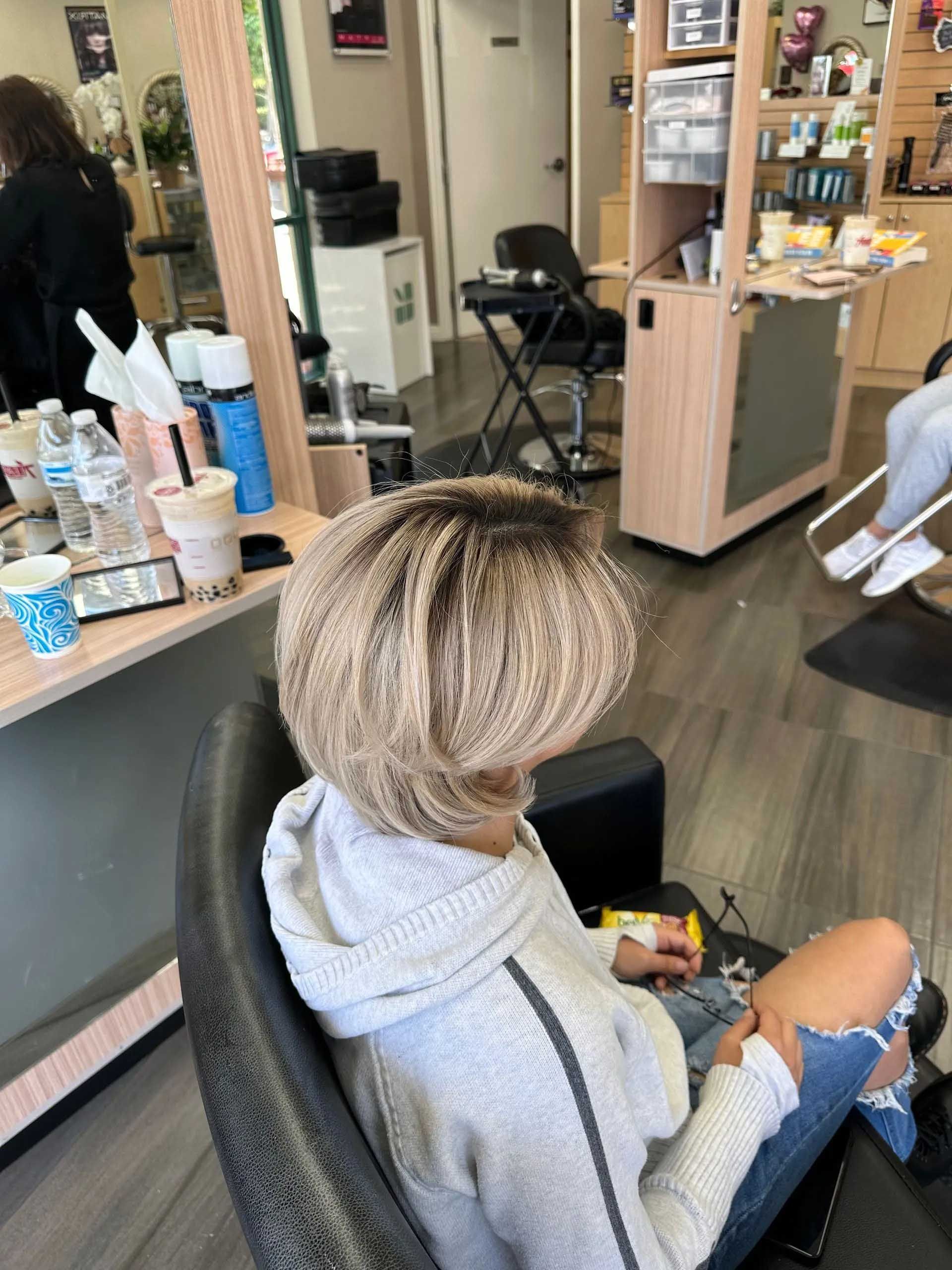 Person in a salon chair with short, layered, light-colored hair, wearing a gray hoodie and ripped jeans.
